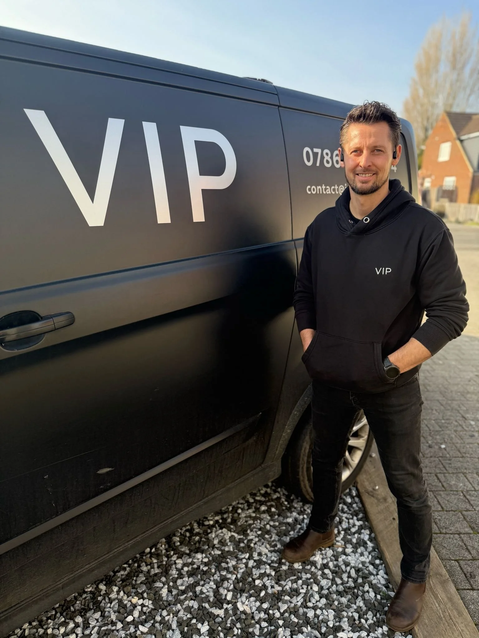 Man standing next to a black vehicle with a large 'VIP' decal on the side, wearing a black hoodie with 'VIP' on it, outdoors on a sunny day.
