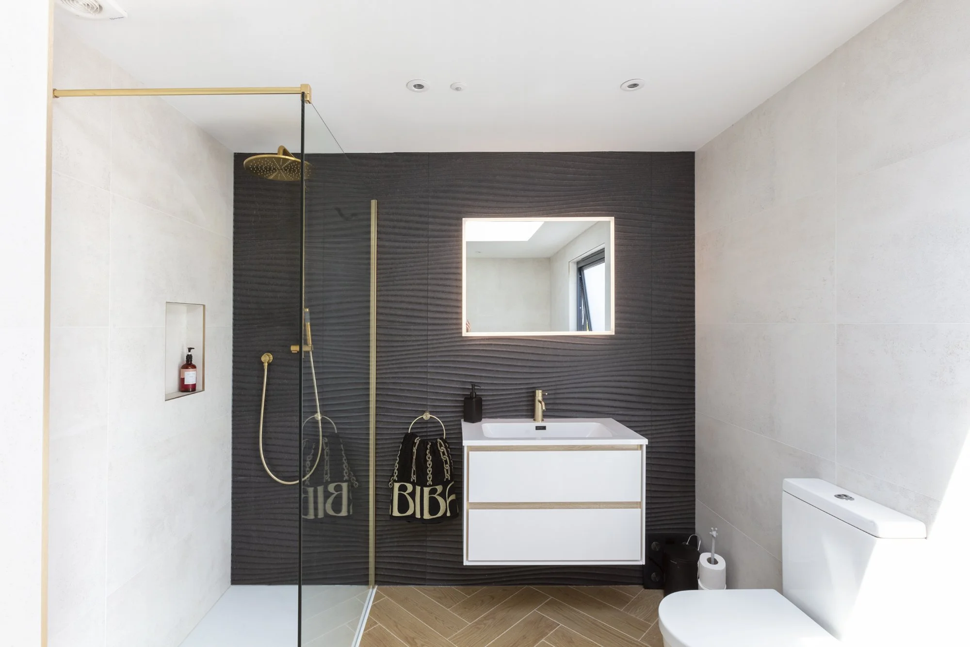 Modern bathroom with black textured wall behind sink and mirror, white fixtures, a shower area with a gold shower head, and toiletries.