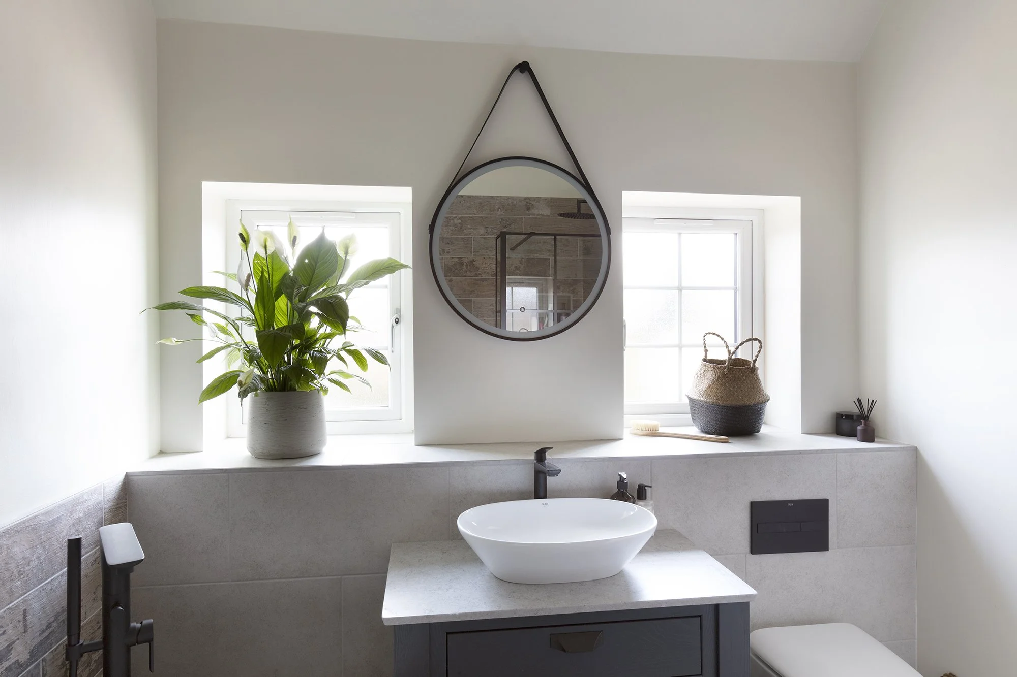 Modern bathroom with a large white vessel sink on a light-colored countertop, a round mirror hanging on the wall, two small windows, a plant in a white pot, woven baskets, and various bathroom accessories.