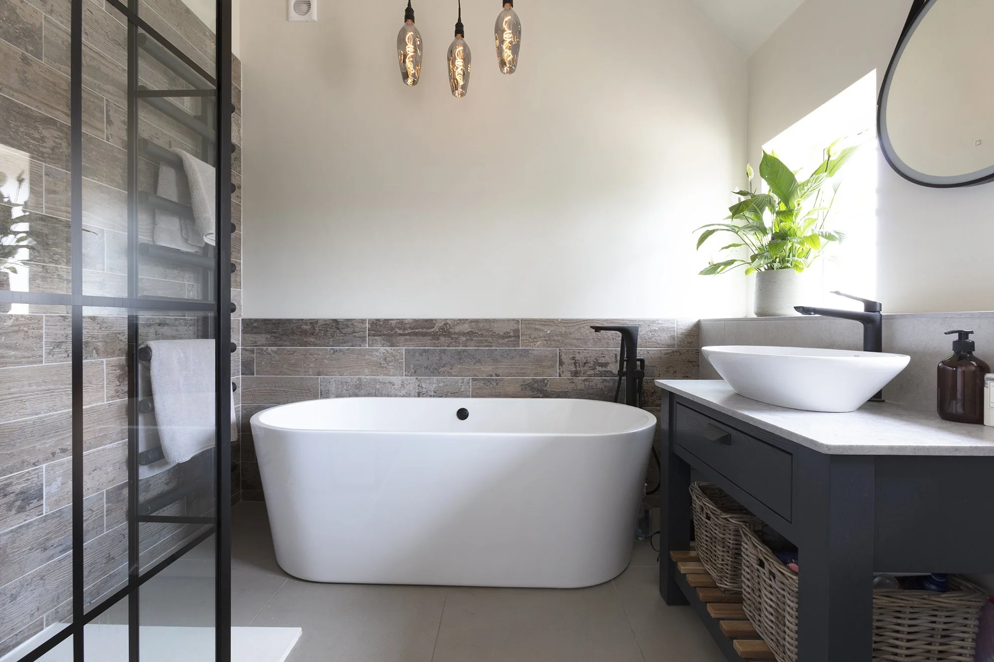 Modern bathroom with a freestanding bathtub, a vanity with a vessel sink, a large green plant in a white pot, and a glass shower enclosure with towels inside. The room has a beige and gray tile design and a minimalist aesthetic, with natural light coming from a window.
