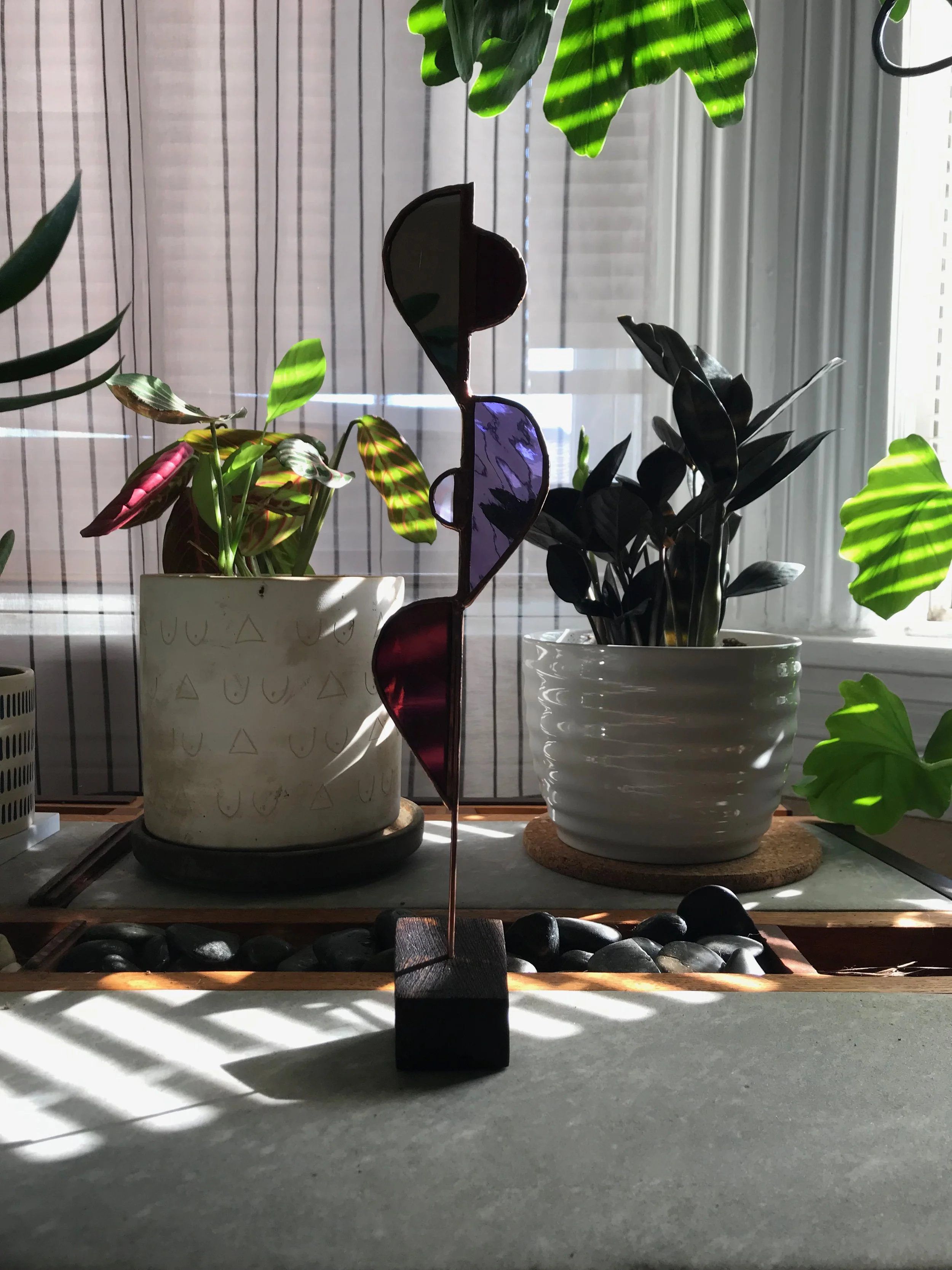Purple, black and dark red freestanding stained glass sculpture with plants in background