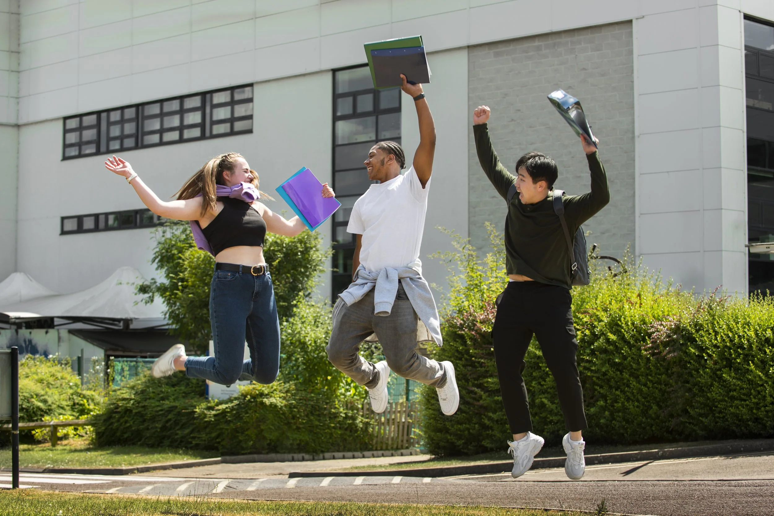 Three young students jumping in celebration outdoors with books and backpacks, standing on a sidewalk in front of a modern building and greenery.