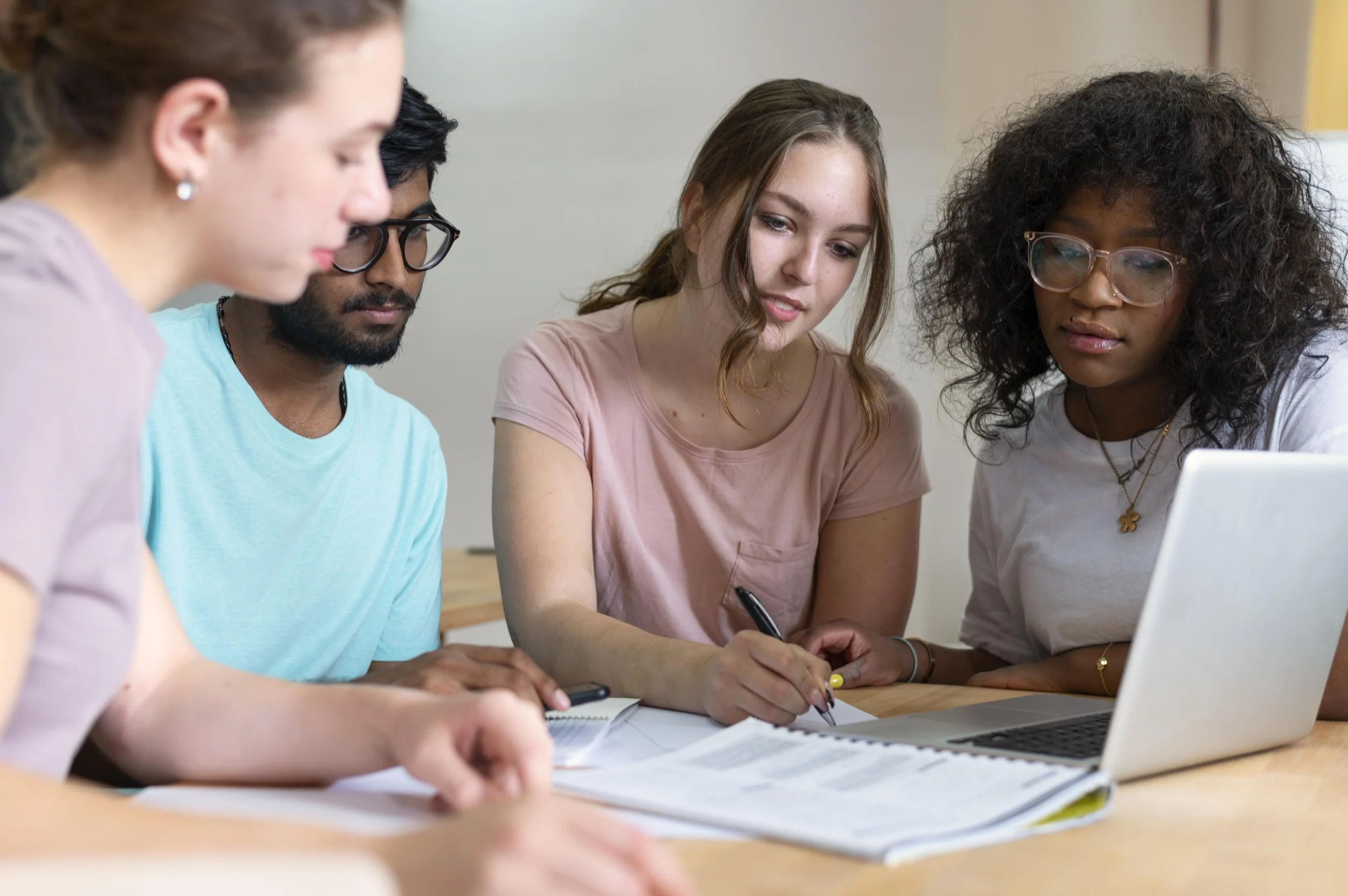 Group of five young adults collaborating at a table, looking at documents and a laptop.