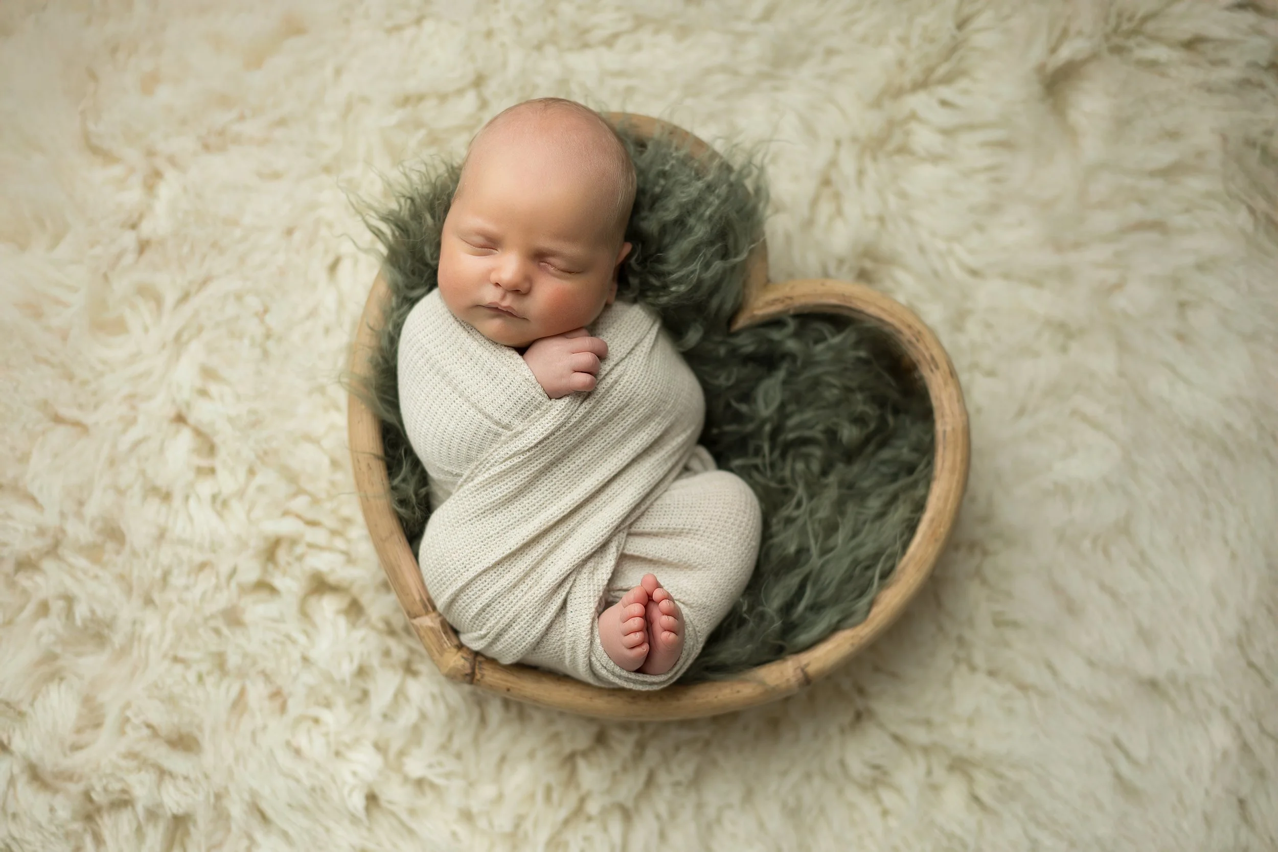Newborn baby sleeping wrapped in a white blanket, placed in a heart-shaped wooden bowl on a fluffy surface.