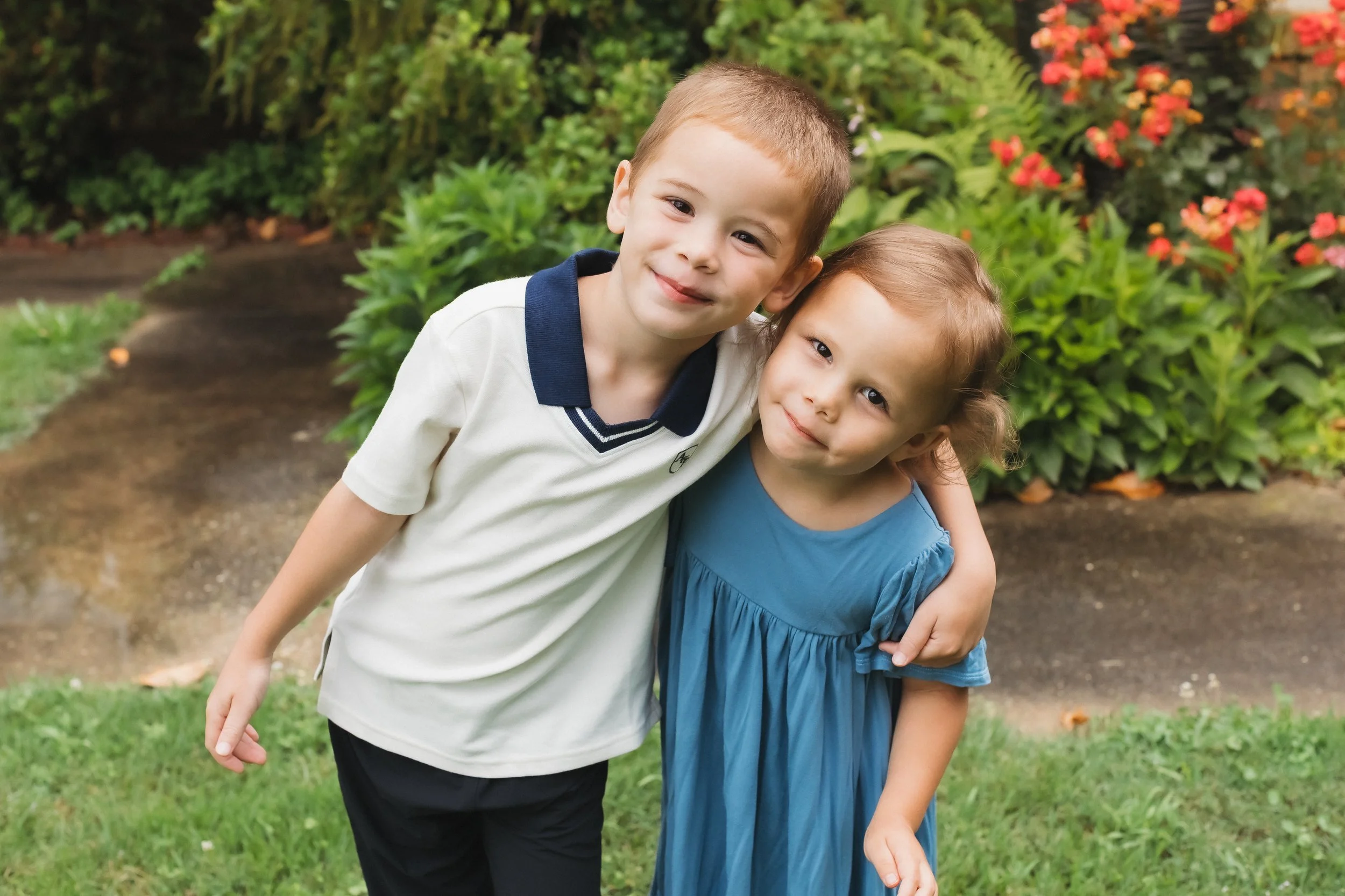 Two children, a boy and a girl, hugging in a garden with green foliage and pink flowers in the background.