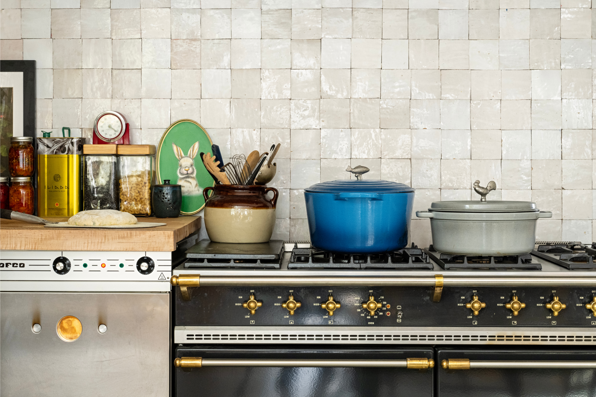 A kitchen counter with canned goods, pasta, black beans, a loaf of bread, cooking utensils in jars, and a stove with a blue and a gray pot.