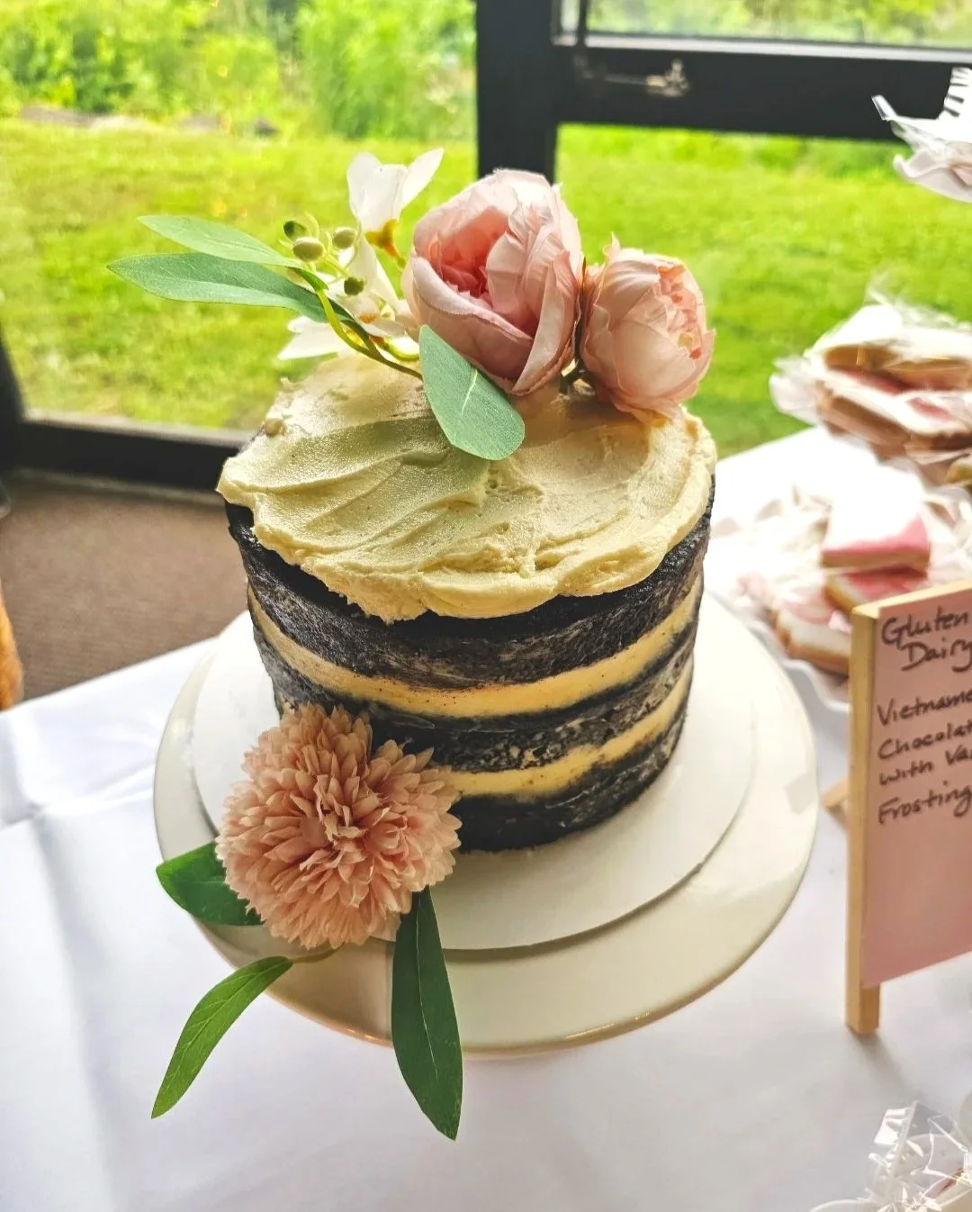 Naked layer cake with Vietnamese coffee cake and vegan condensed milk frosting, decorated with pink silk flowers and green leaves, on a white cake stand. Custom gluten free and dairy free cake.