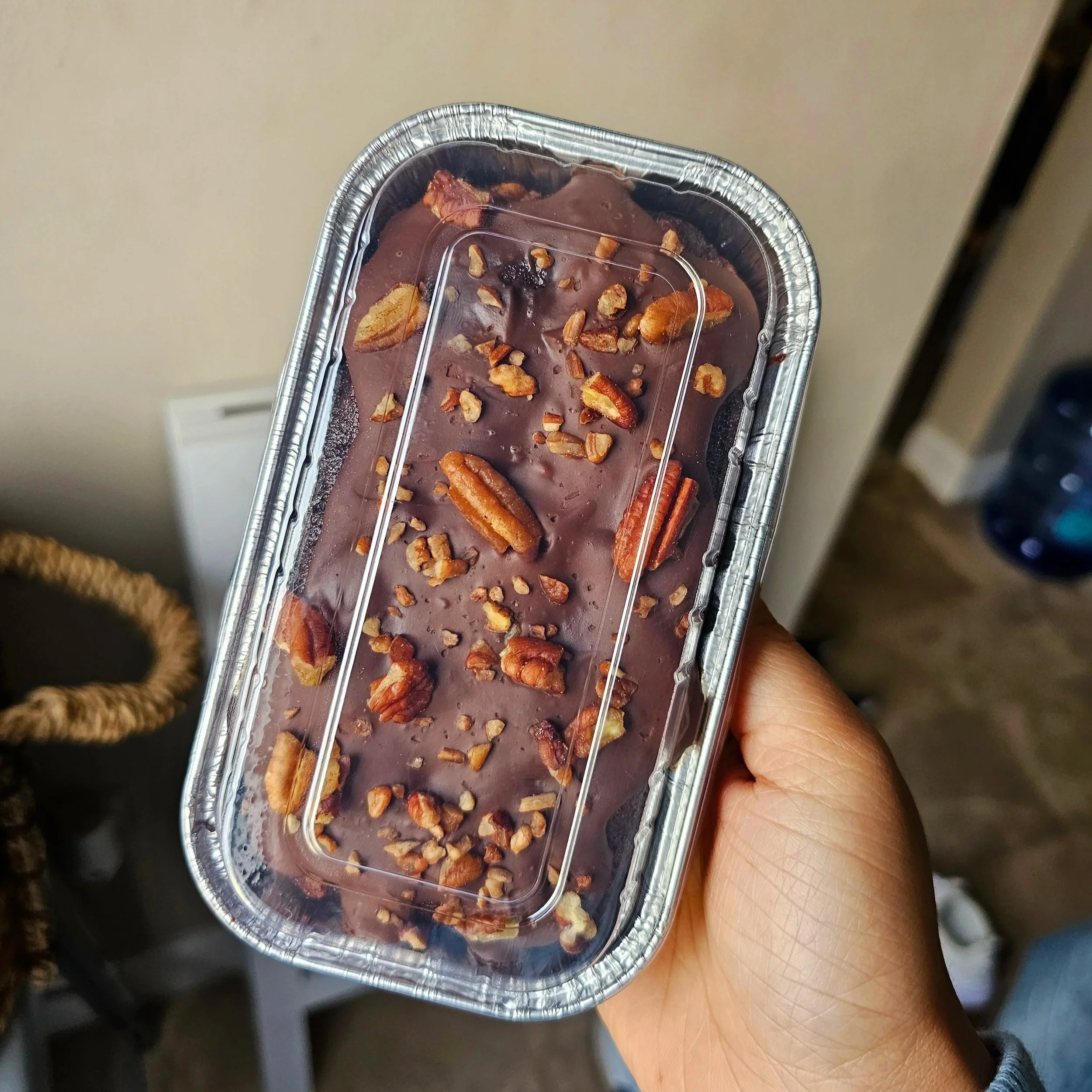 A hand holding a foil container with chocolate cake dessert topped with pecans and vegan fudge glaze