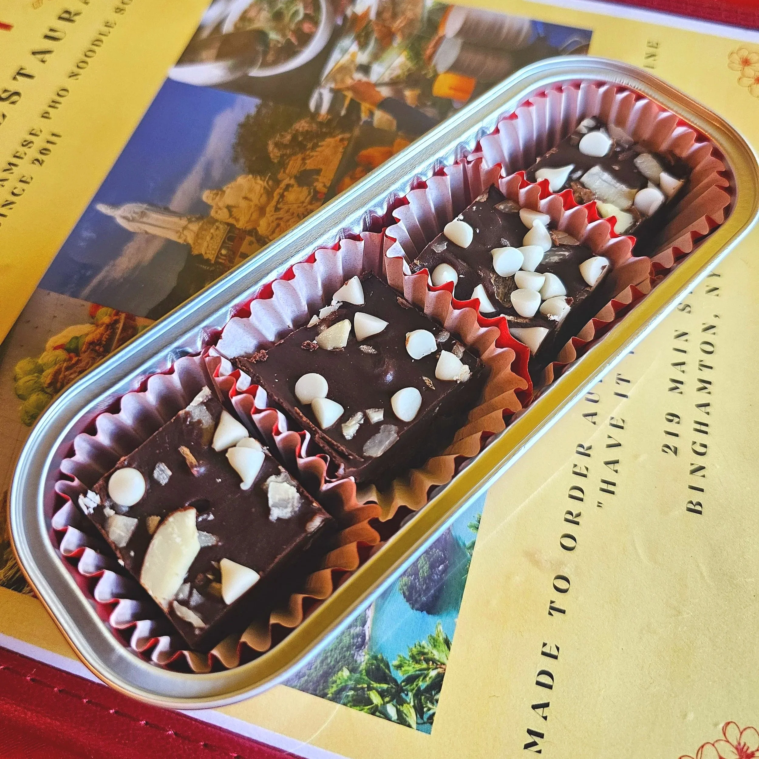Four pieces of vegan fudge topped with white chocolate chips and almonds, placed in a red and white paper-lined metal container on a Vietnamese restaurant menu. Dairy Free Vietnamese coffee fudge