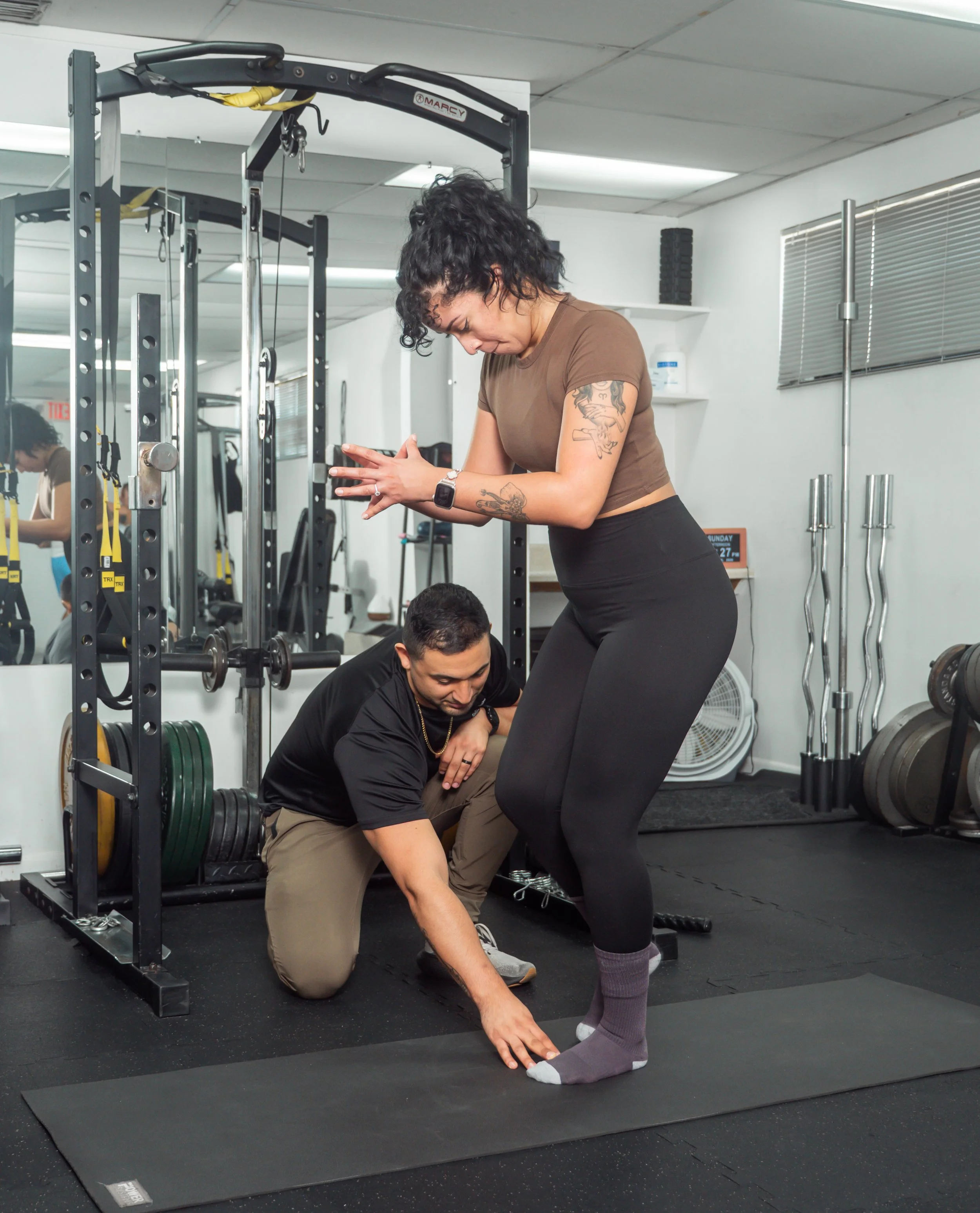 A woman with tattoos and curly hair practicing yoga while a man assists her in a gym. The gym has weightlifting equipment and mirrors.