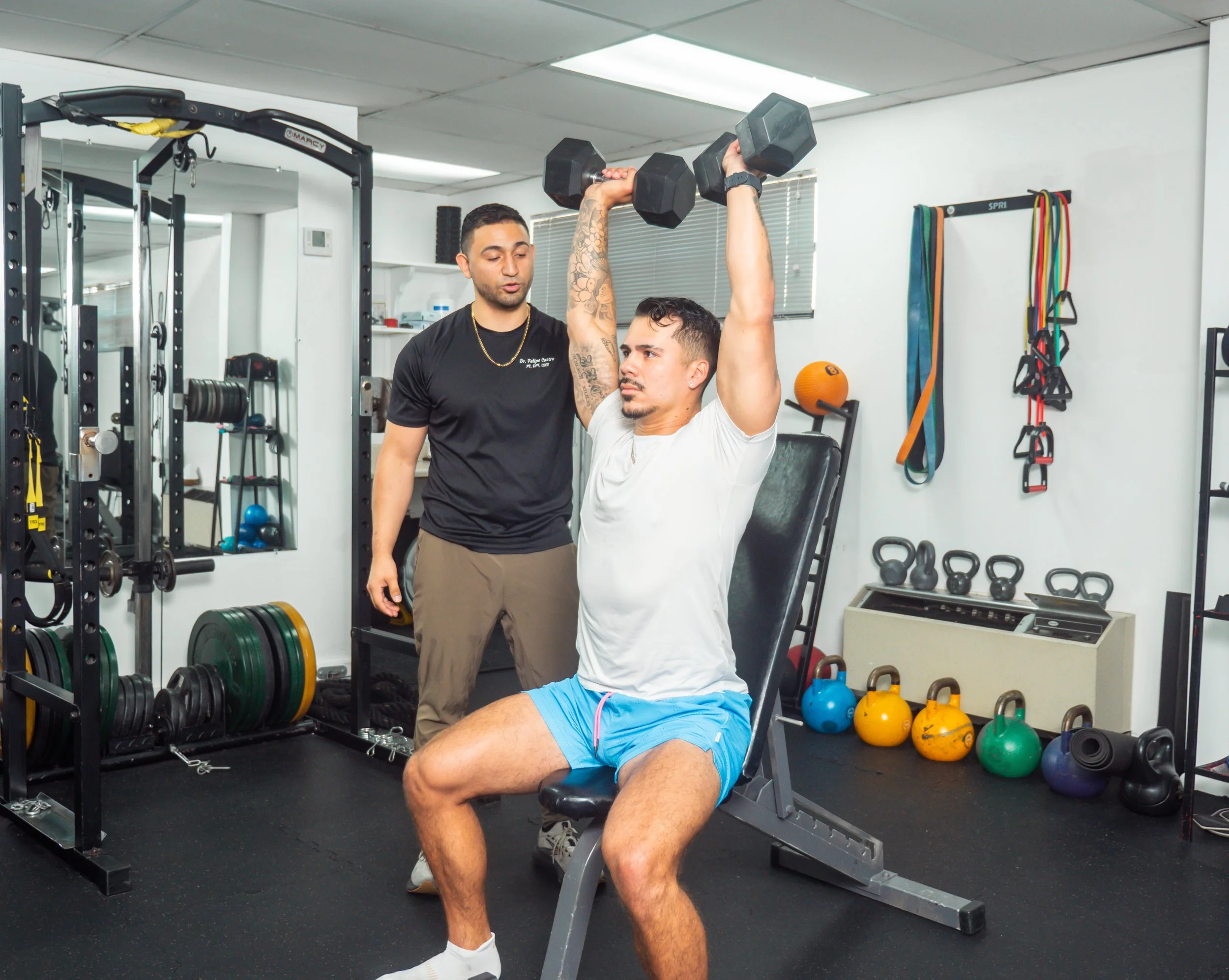 A man in a white t-shirt and blue shorts performs a shoulder press with dumbbells while sitting on a workout bench, under the supervision of a personal trainer in a gym.