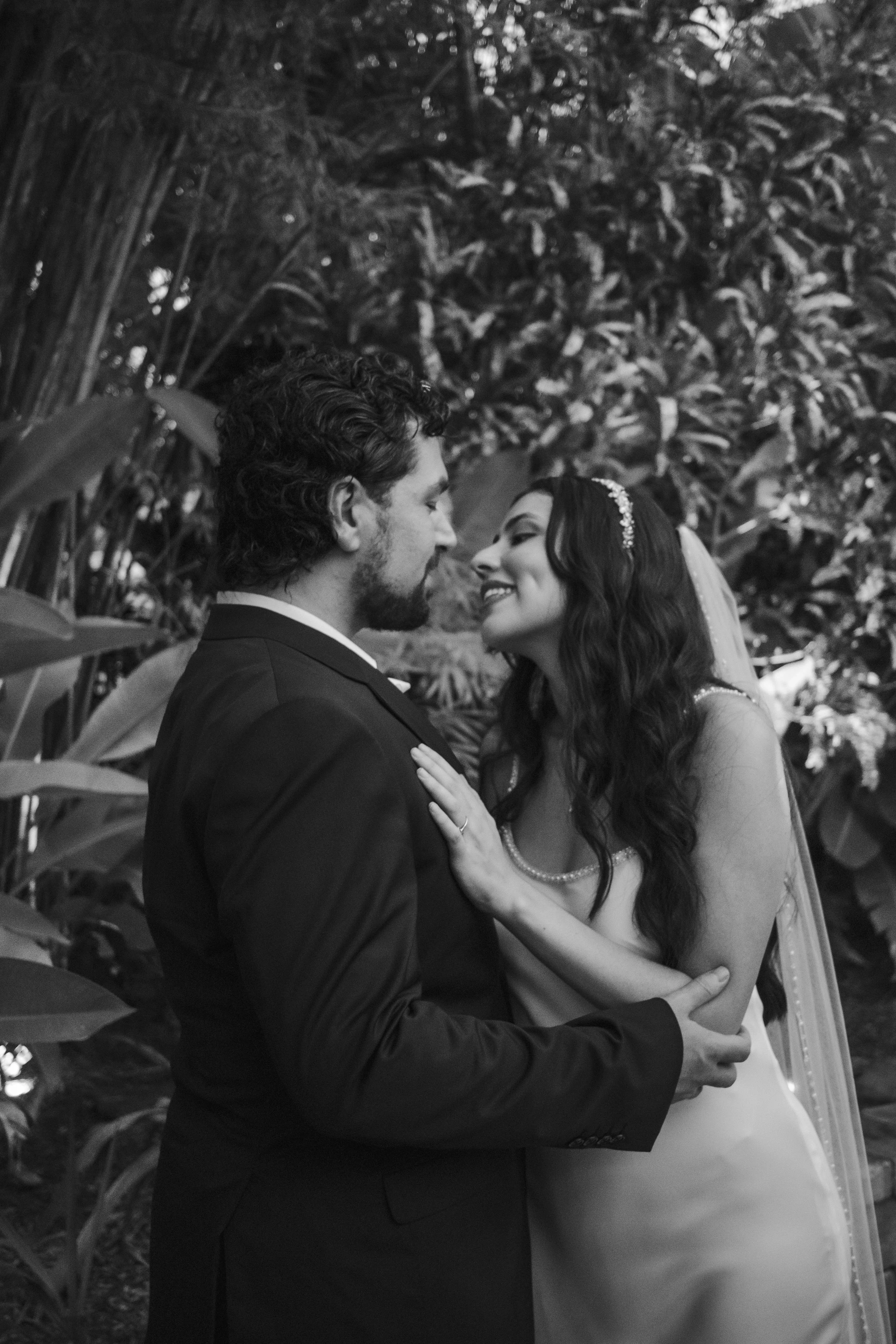A couple dressed in wedding attire holding each other and looking into each other's eyes surrounded by lush foliage.