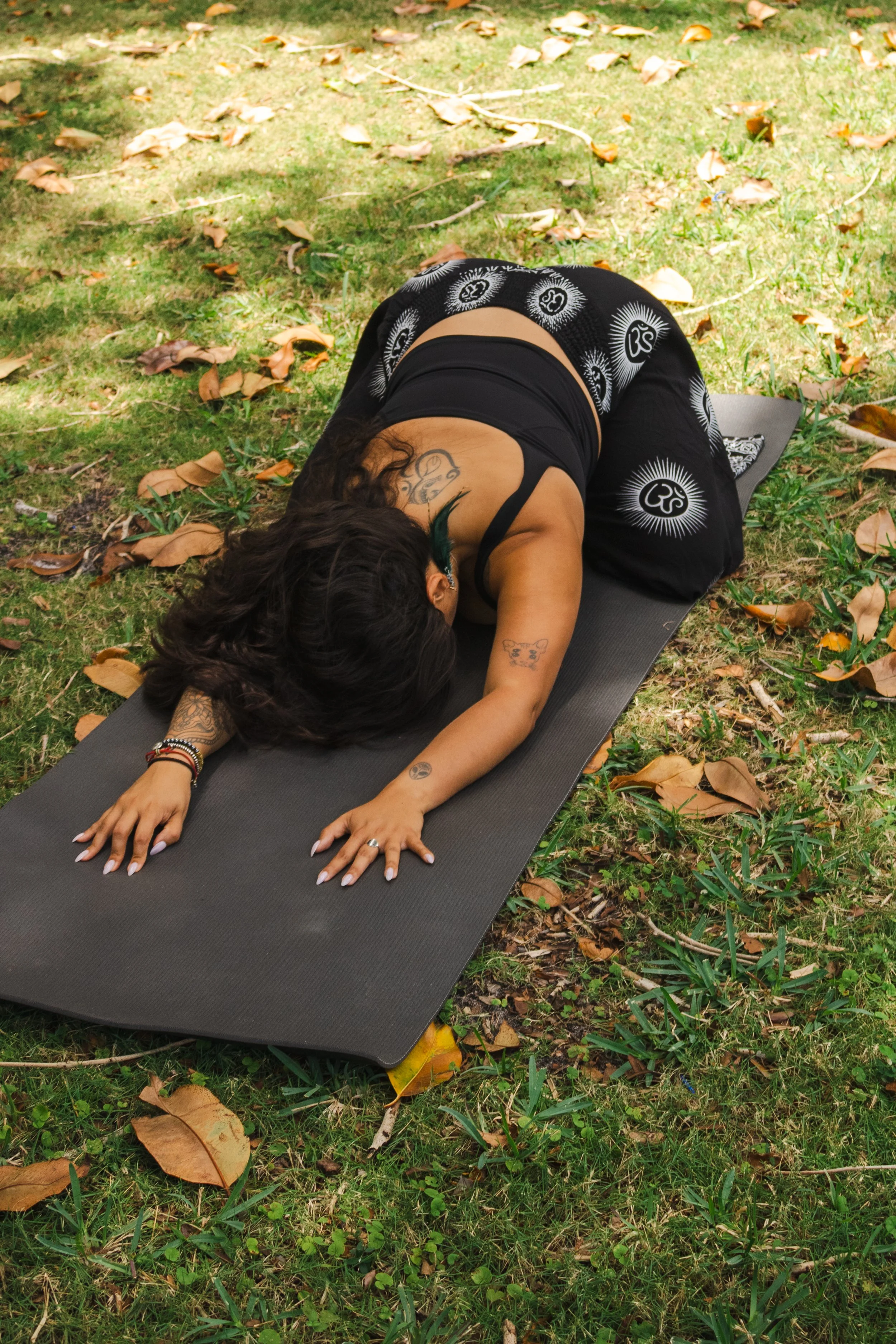 A person is practicing yoga outdoors on a black yoga mat, laying in child's pose with arms extended forward, surrounded by grass and fallen leaves.