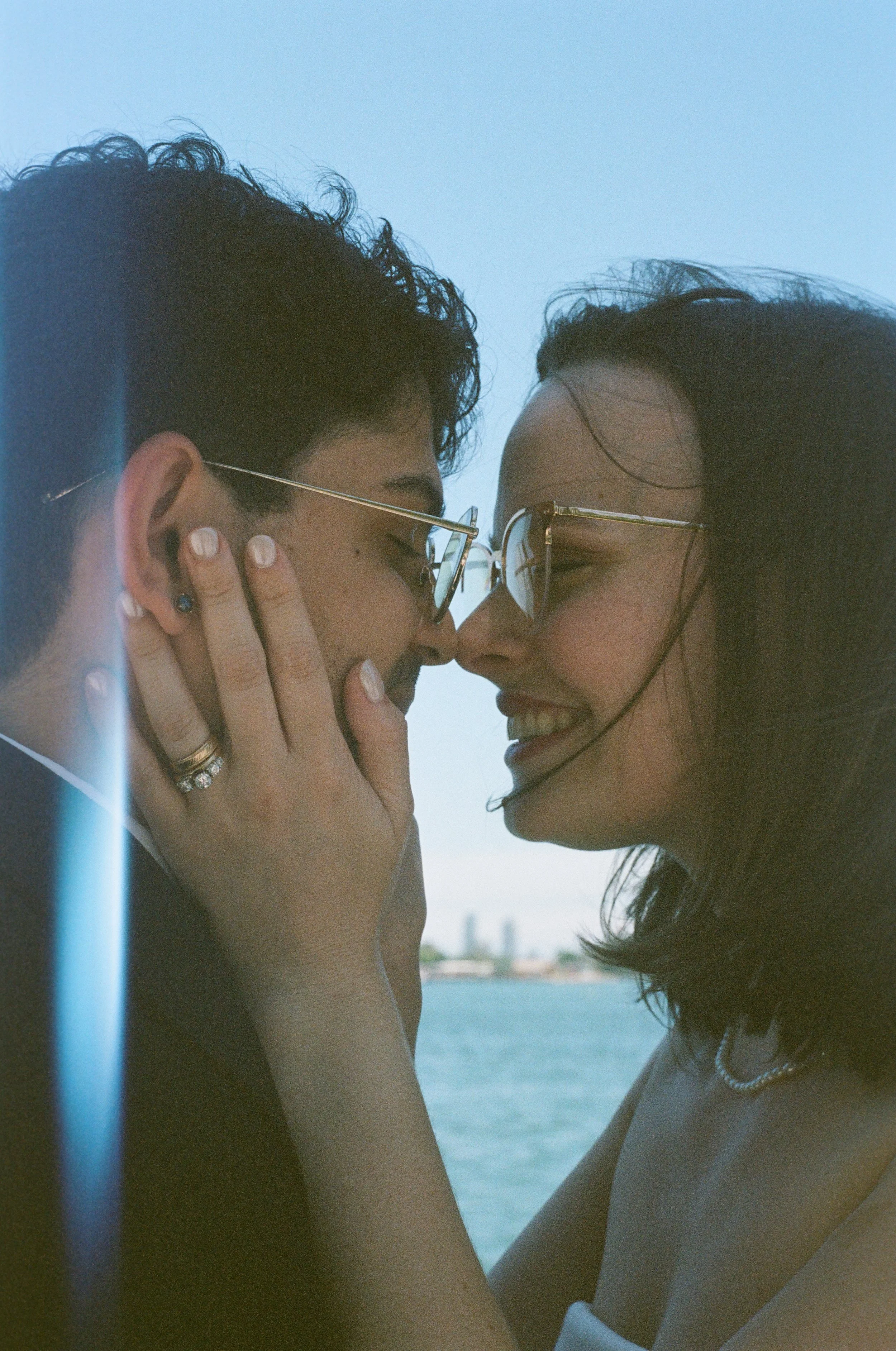 A close-up of a couple with glasses sharing an intimate moment near a body of water, with city skyline in the background.