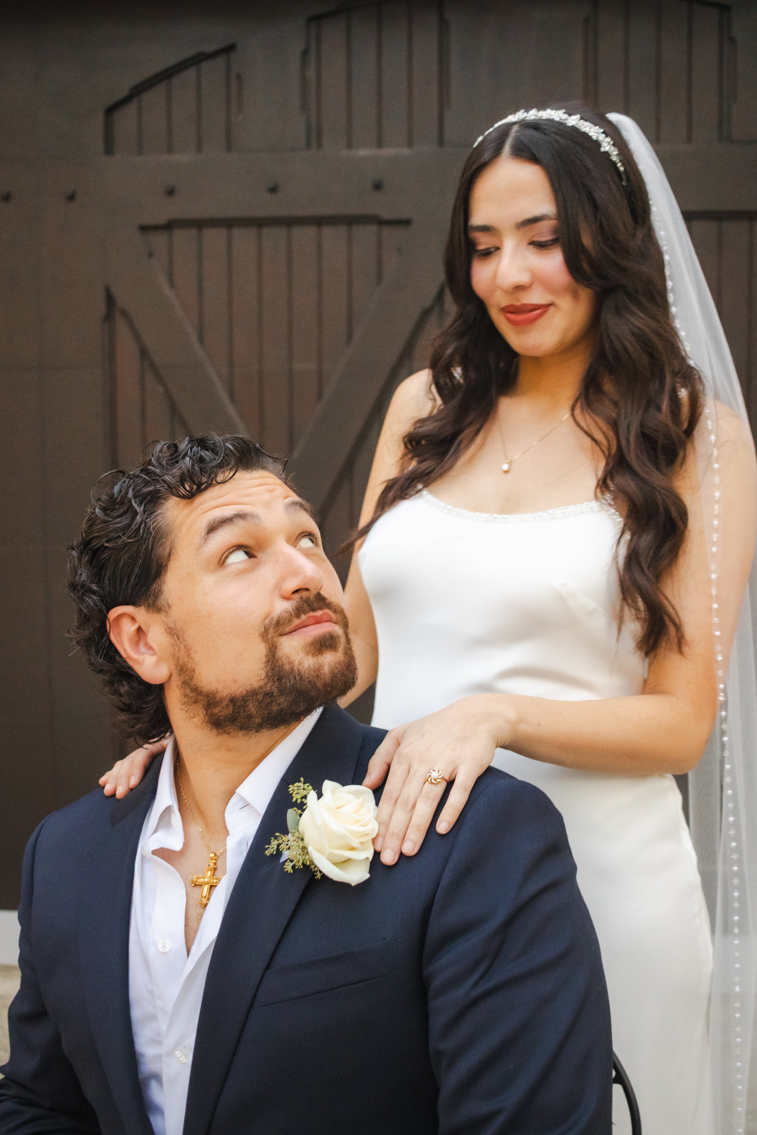 A woman with long dark hair dressed in a white bridal gown and a veil with a beaded headband, looking down with a gentle smile. A man dressed in a dark suit with a white shirt, sporting a beard and curly hair, sits in front of her, looking up at her 
