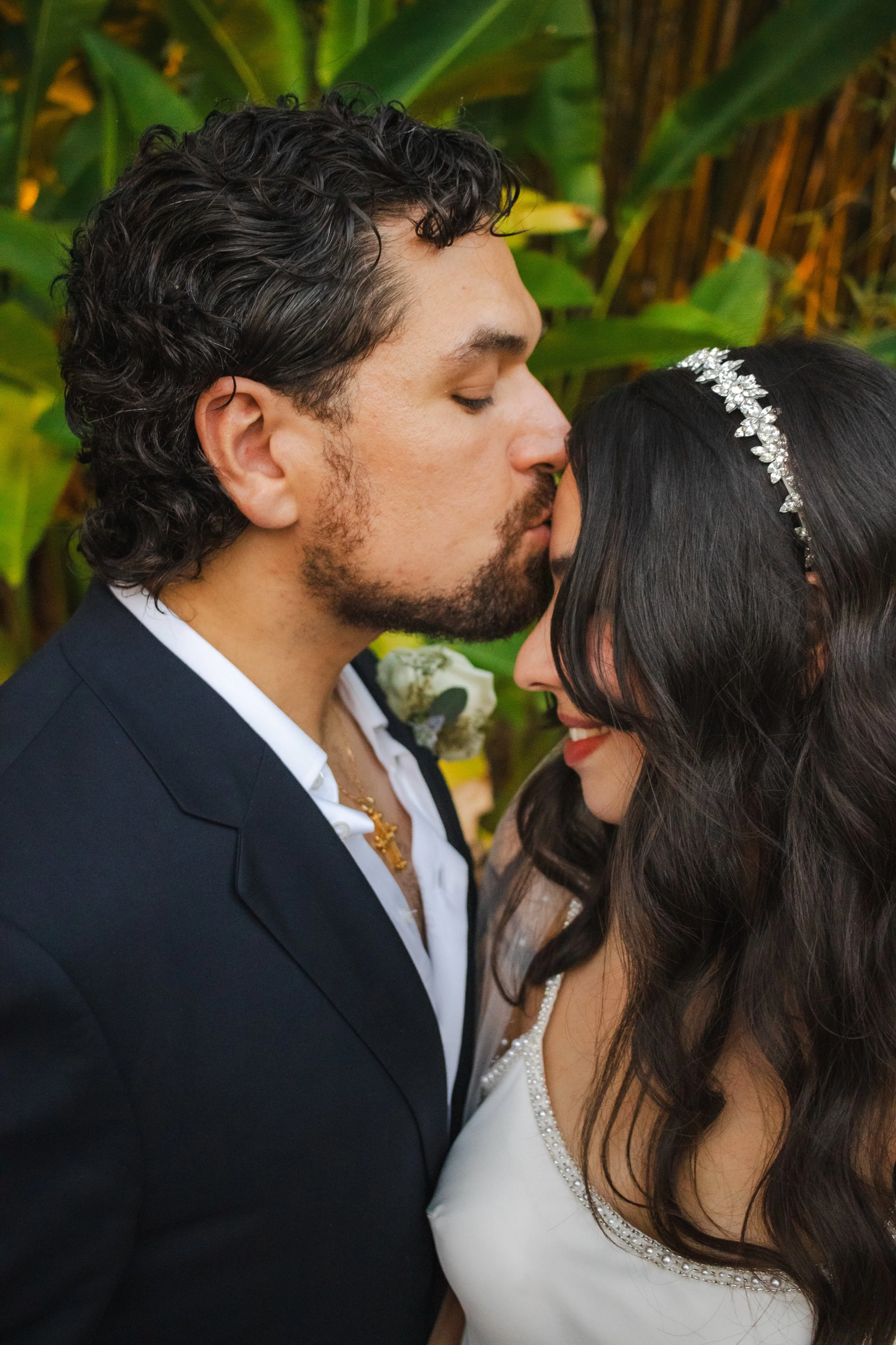 A couple dressed in wedding attire sharing an intimate moment, with the man in a dark suit and the woman in a white wedding dress with pearl embellishments, surrounded by greenery.