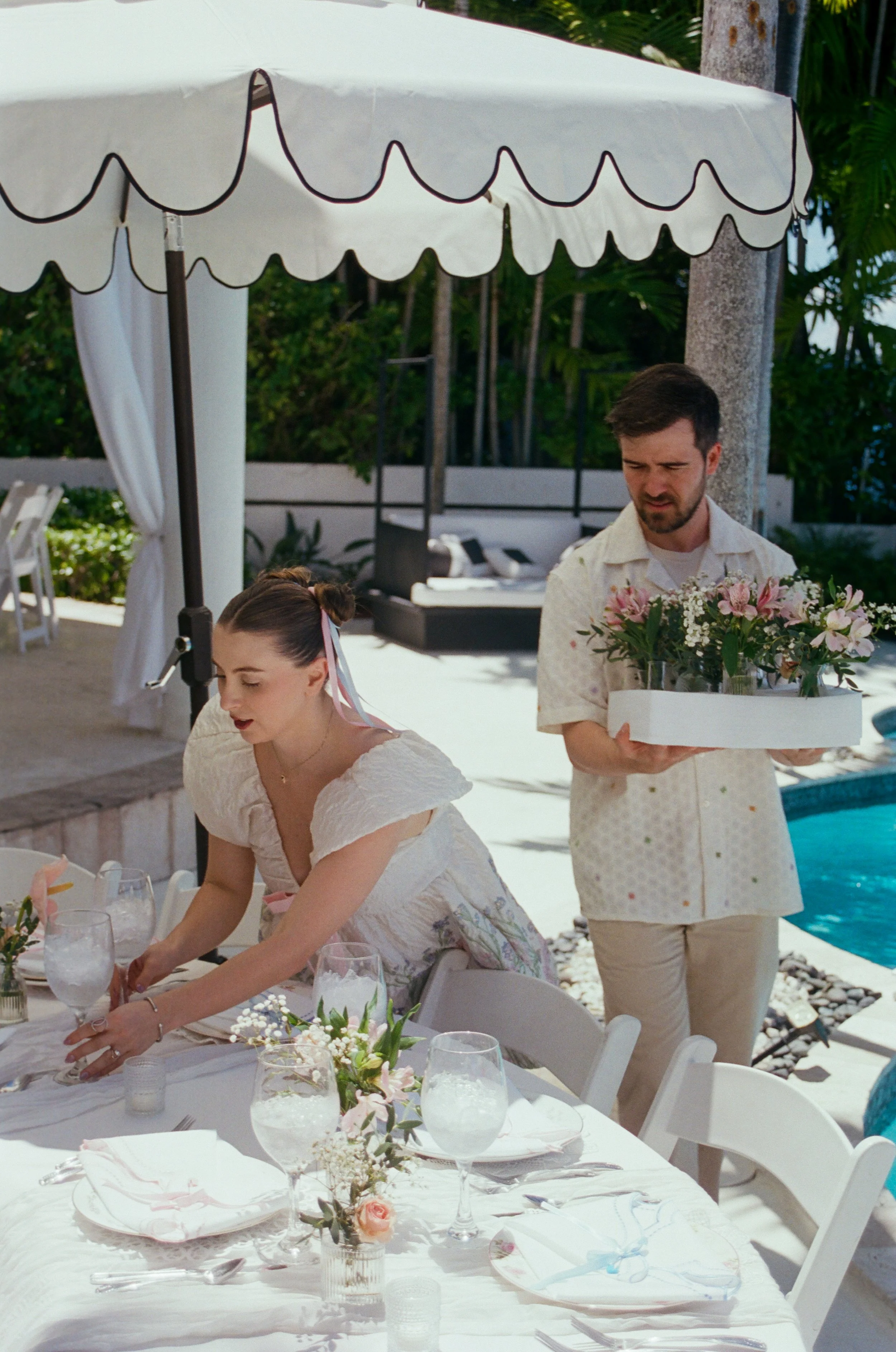 People setting a table outdoors under a white canopy, with floral arrangements and place settings visible, near a pool and surrounded by trees.