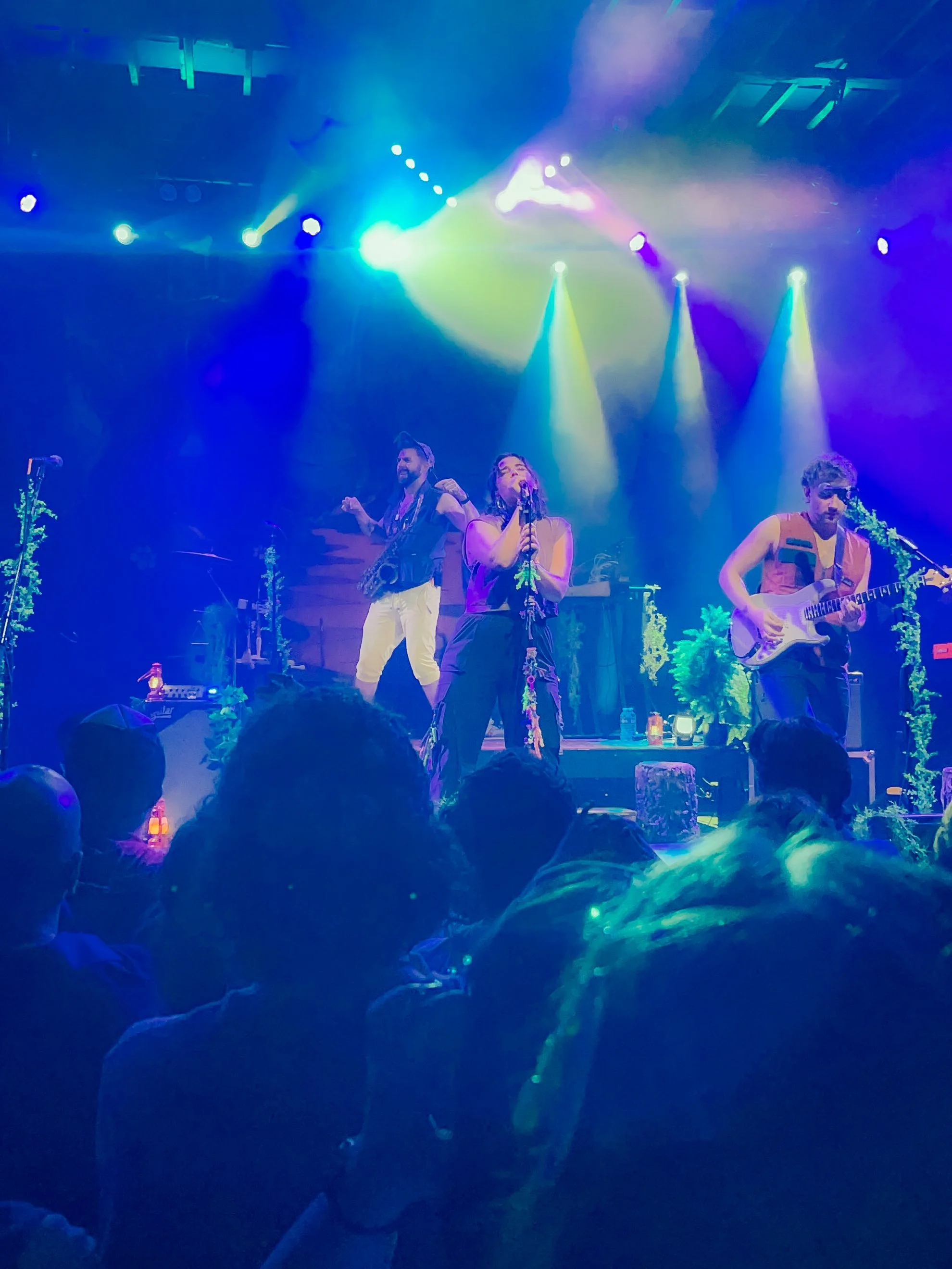 A live music performance with three musicians on stage illuminated by colorful stage lights, with an audience in front.