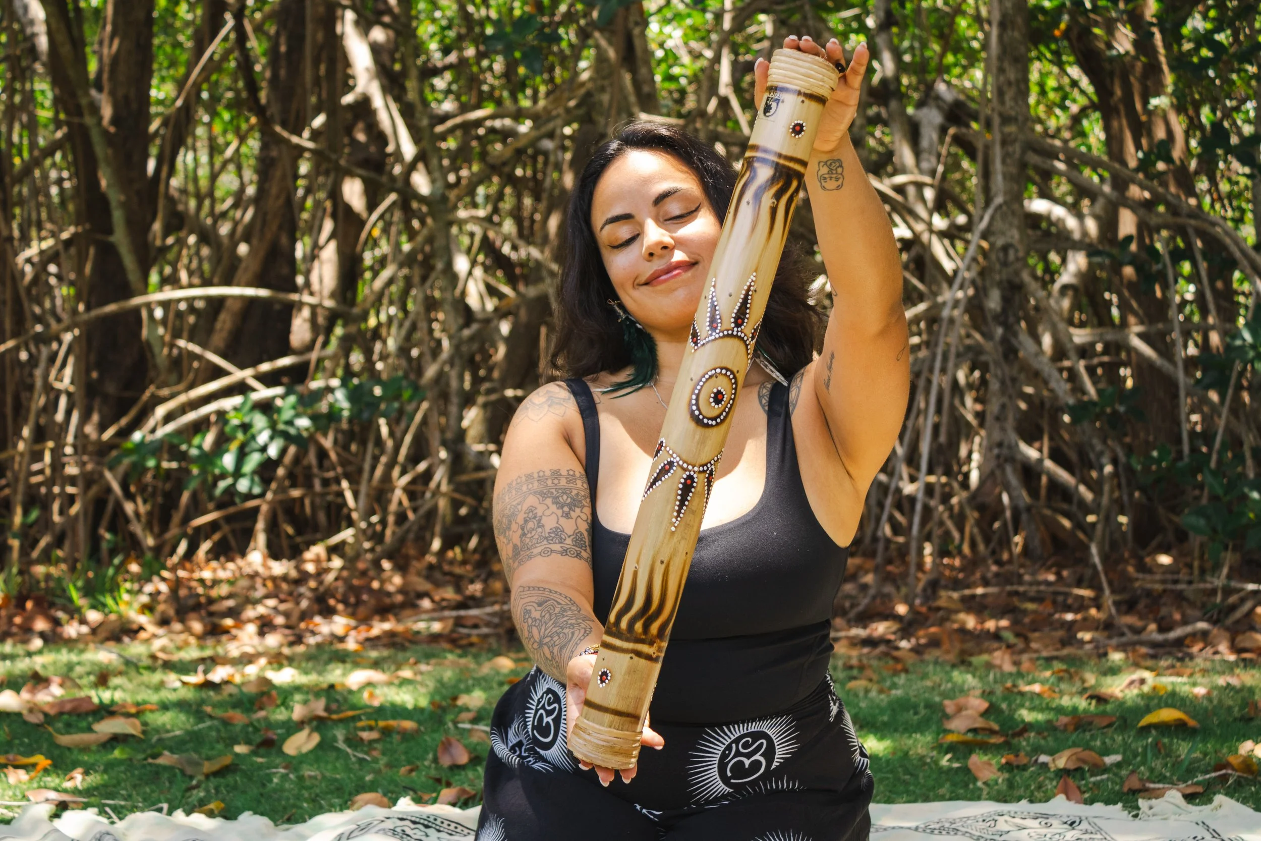 A woman with tattoos on her arms and shoulder, wearing a black tank top and patterned pants, stands in a wooded outdoor area holding a decorated bamboo flute. She appears content with closed eyes and a gentle smile on her face.