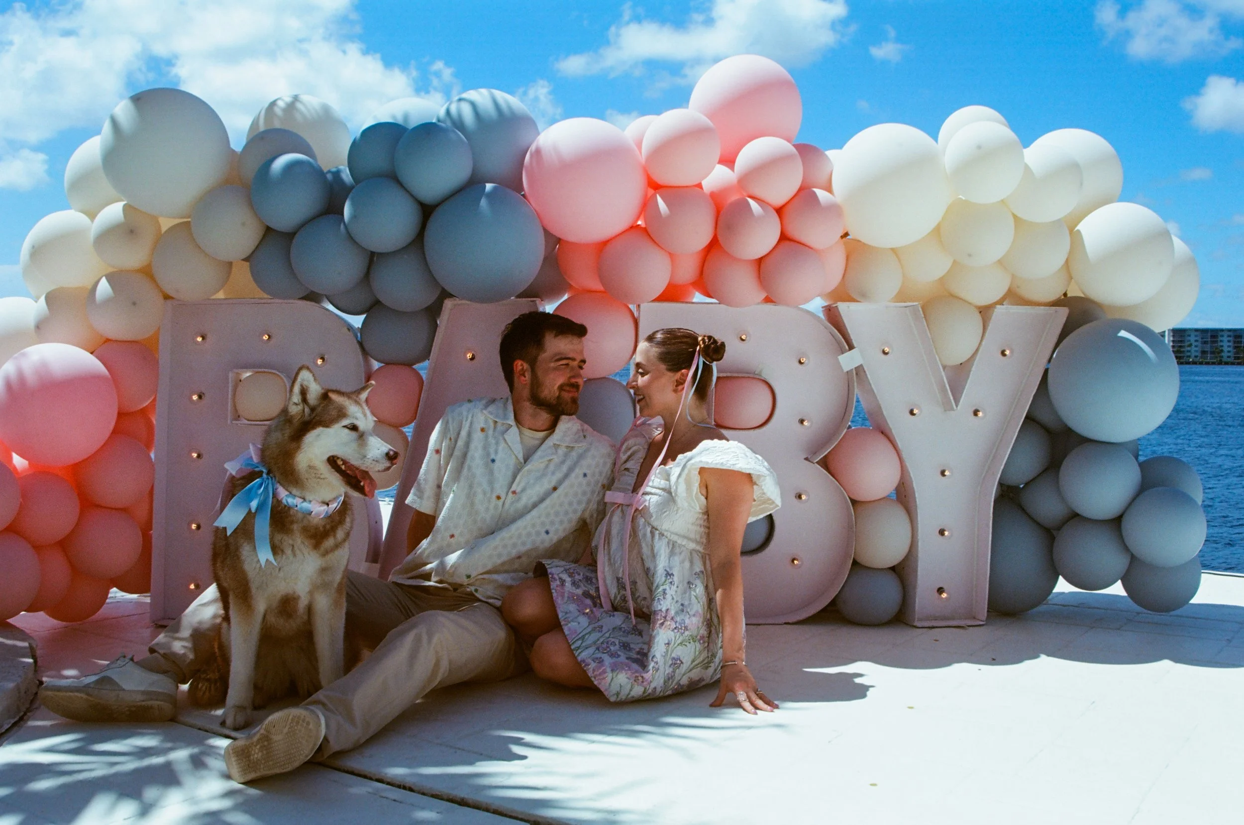 A couple sitting on the ground in front of a decorated balloon display with a dog beside them. The display spells out the word 'LOVE' with large illuminated letters and is adorned with pink, white, and blue balloons. The setting appears to be outdoor