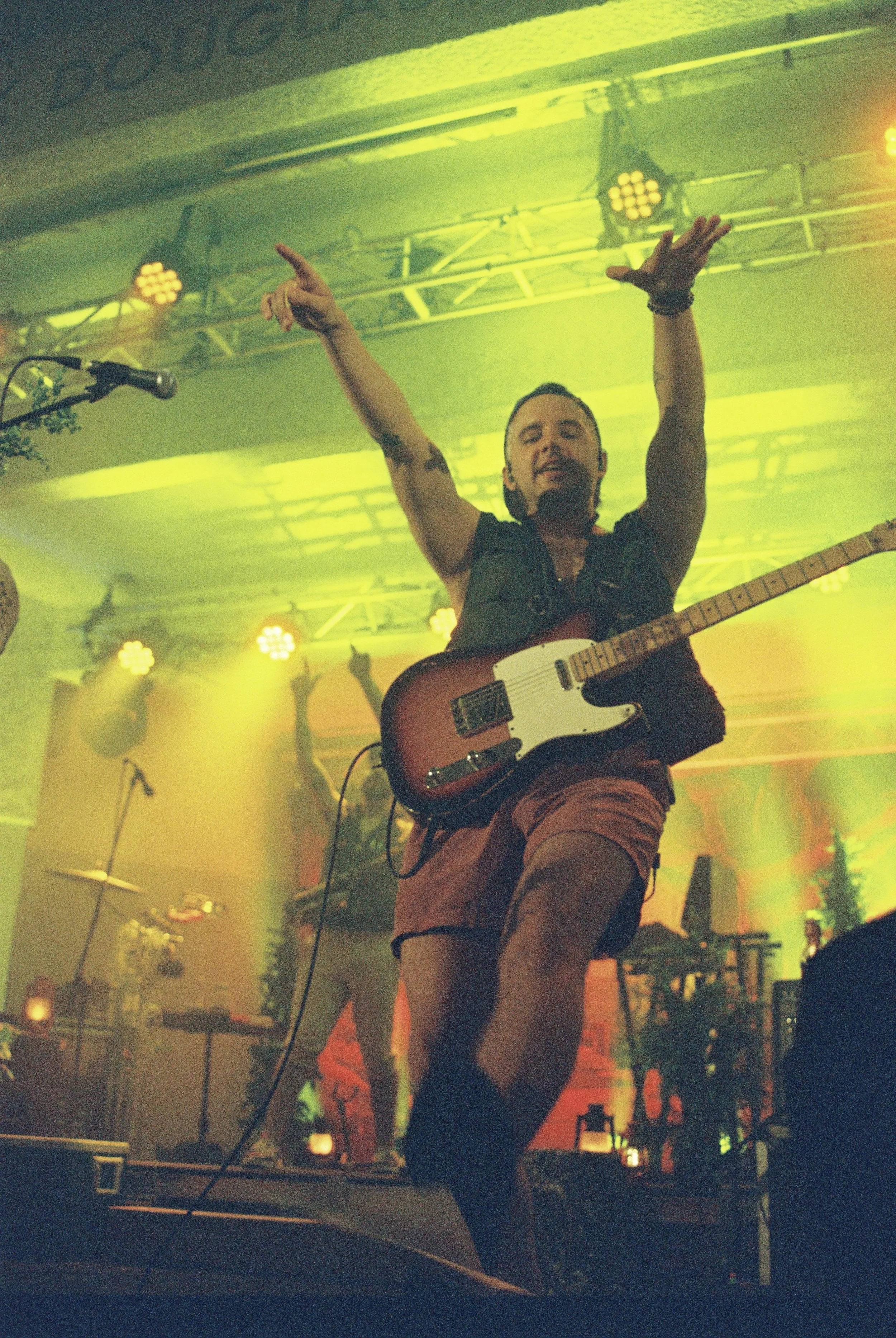 A man with short hair and a beard, playing an electric guitar on stage. He is smiling, has his arms raised, and is wearing a sleeveless dark shirt and shorts. Bright yellow and green stage lights illuminate the background with musical equipment and f
