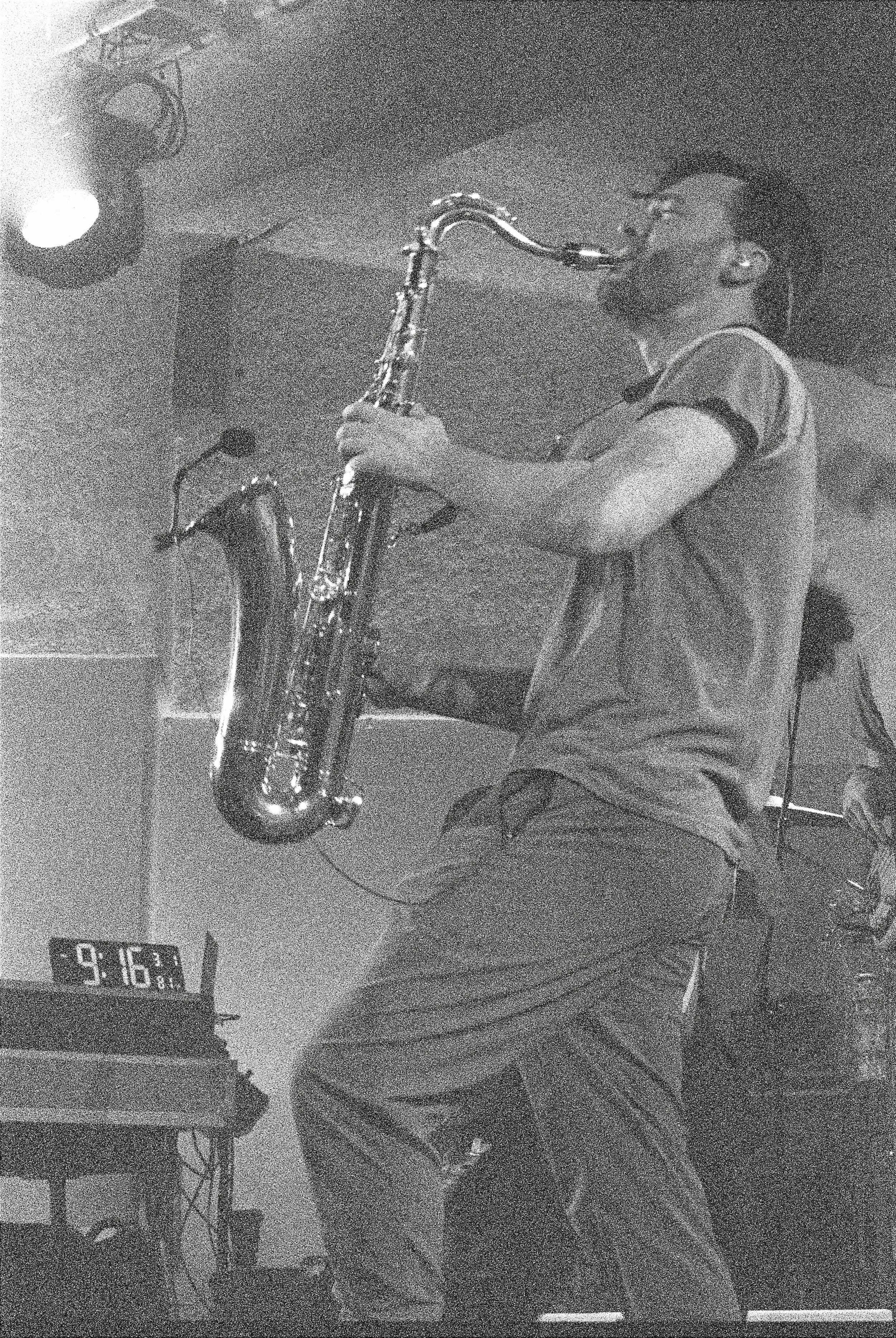 A person playing a saxophone on a stage, there is a clock showing 9:16 as a reminder of their time remaining. The person appears to be wearing a casual T-shirt.