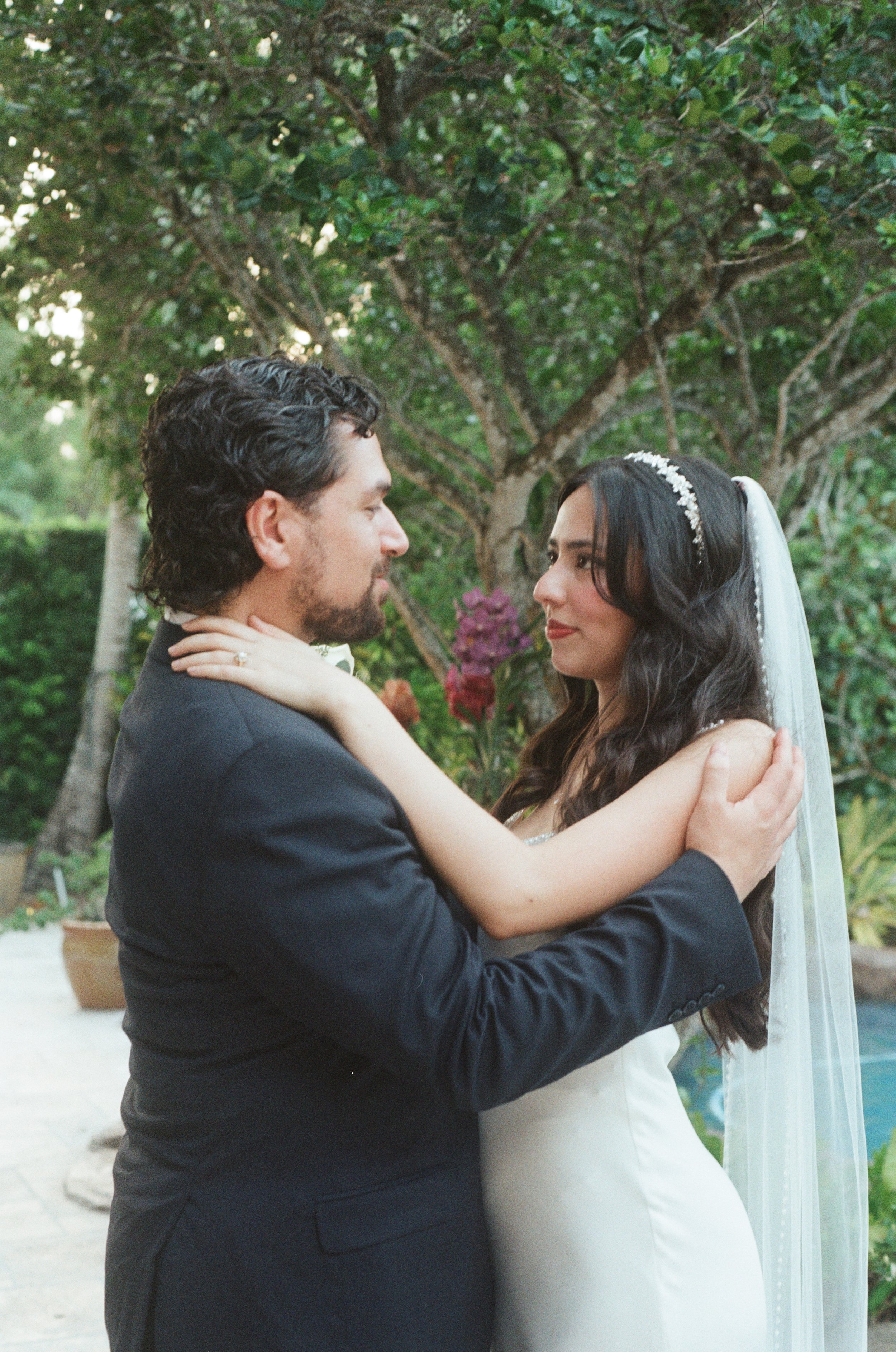 A bride and groom holding each other and looking into each other's eyes outdoors with greenery in the background.