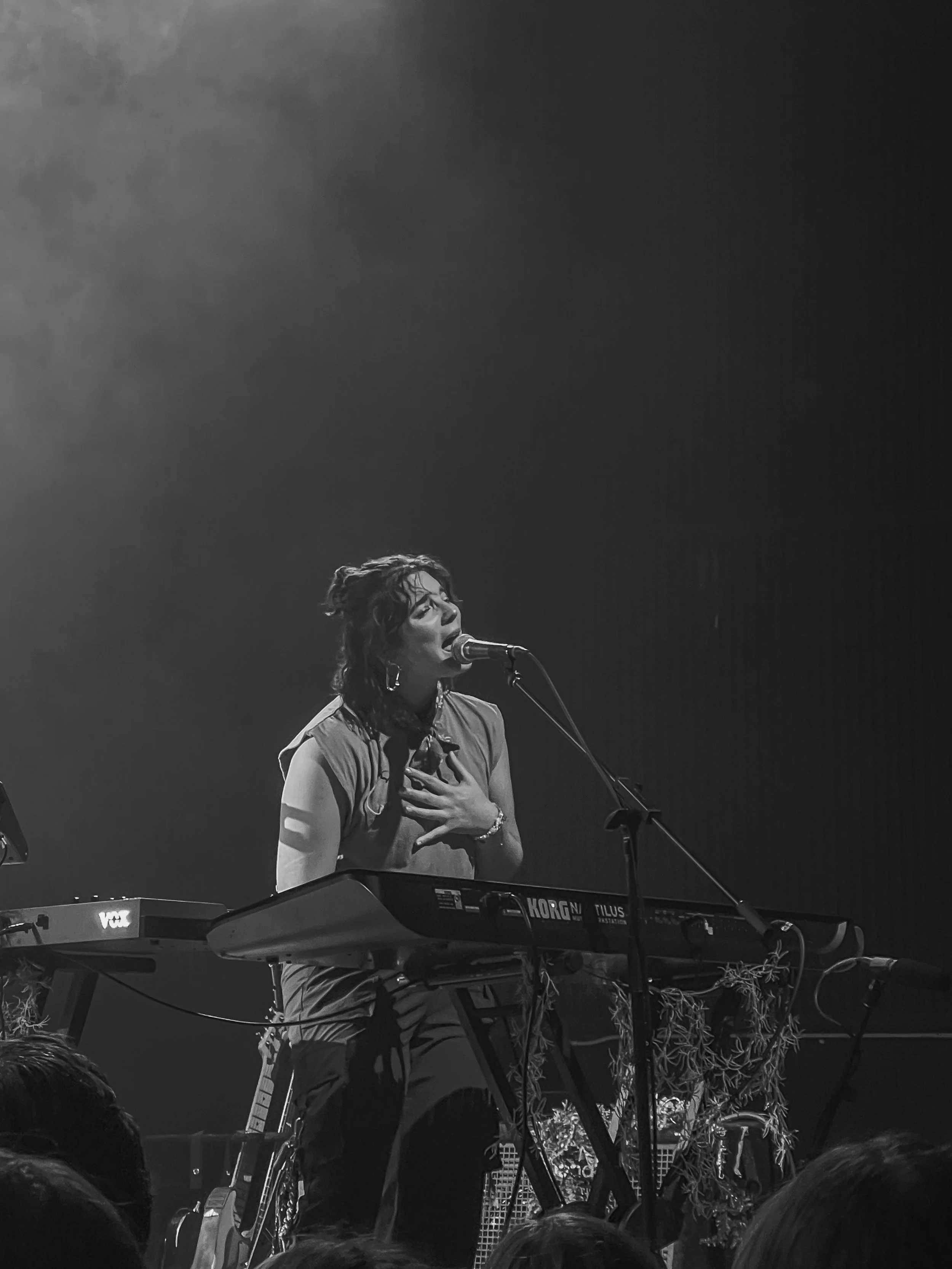 A black and white photo of a woman singing passionately into a microphone on a stage, standing behind a keyboard with the brand 'Korg' visible.
