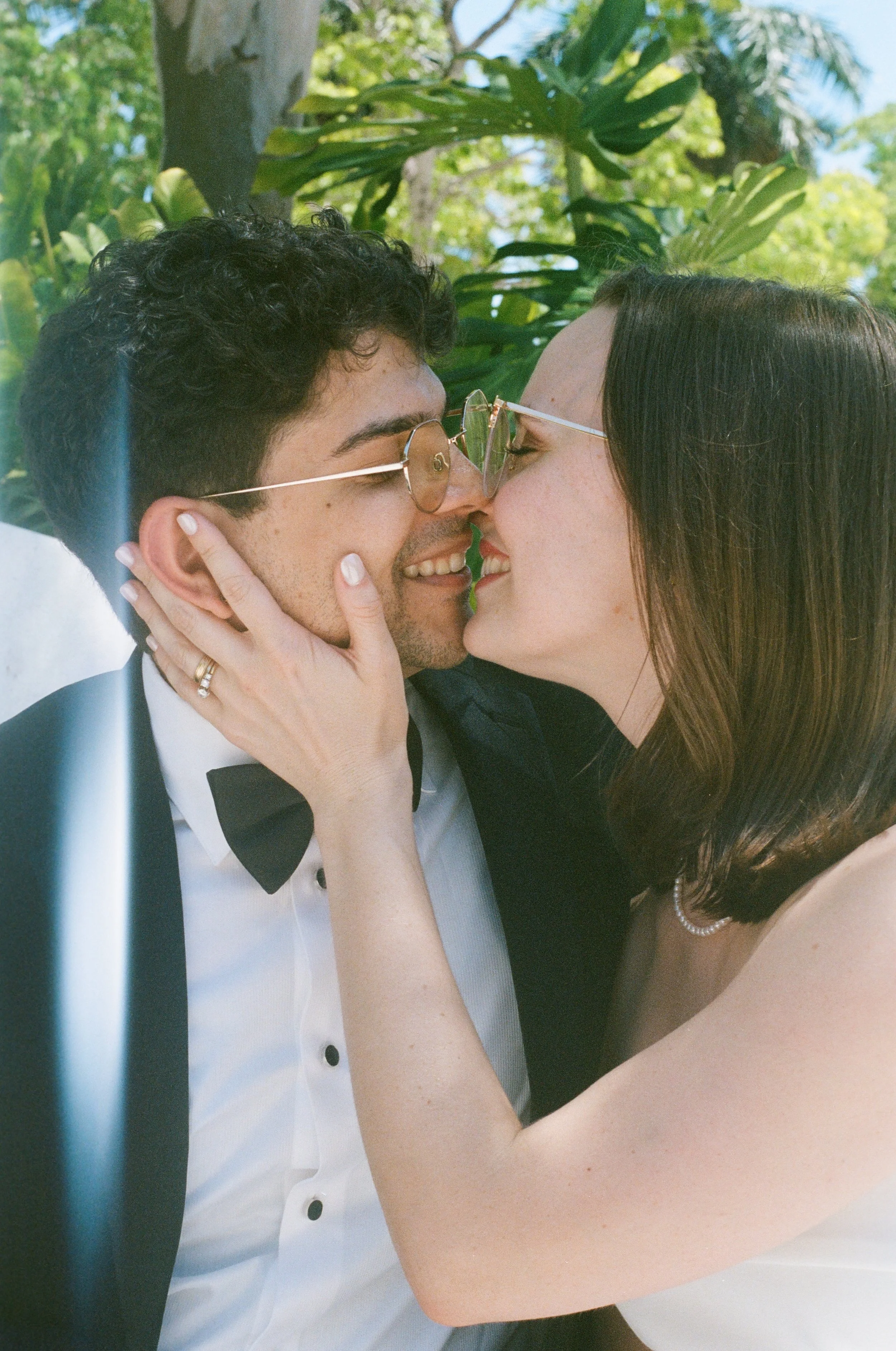 A couple kissing outdoors with trees and blue sky in the background. The woman has long brown hair and is wearing a pearl necklace and a sleeveless dress, while the man has short , curly hair, glasses, and is wearing a tuxedo with a bow tie. The woma