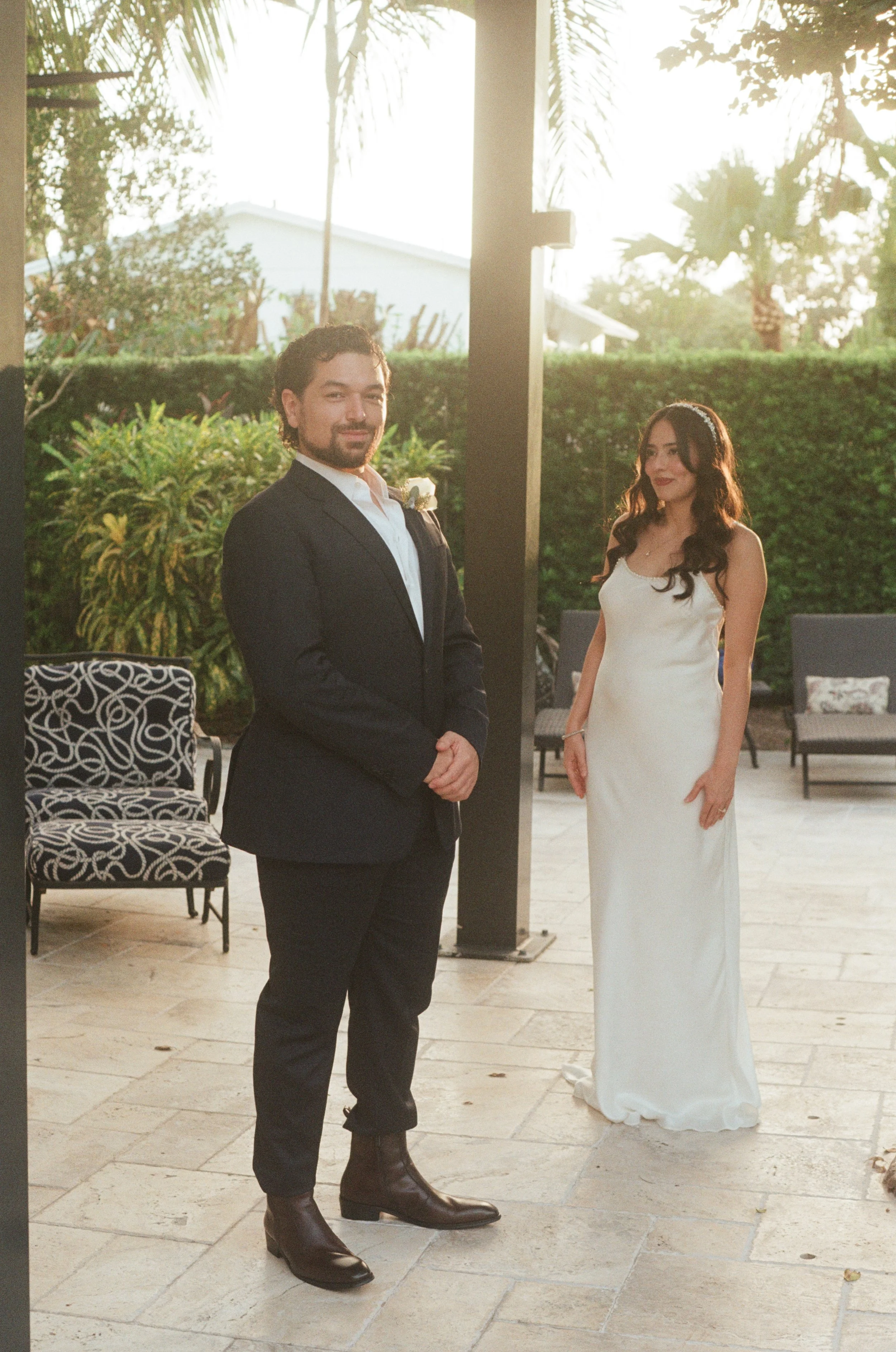 A man in a black suit, white shirt, and dark boots stands outdoors near a woman in a white dress. The background features greenery, including trees and bushes, with sunlight creating a warm glow.