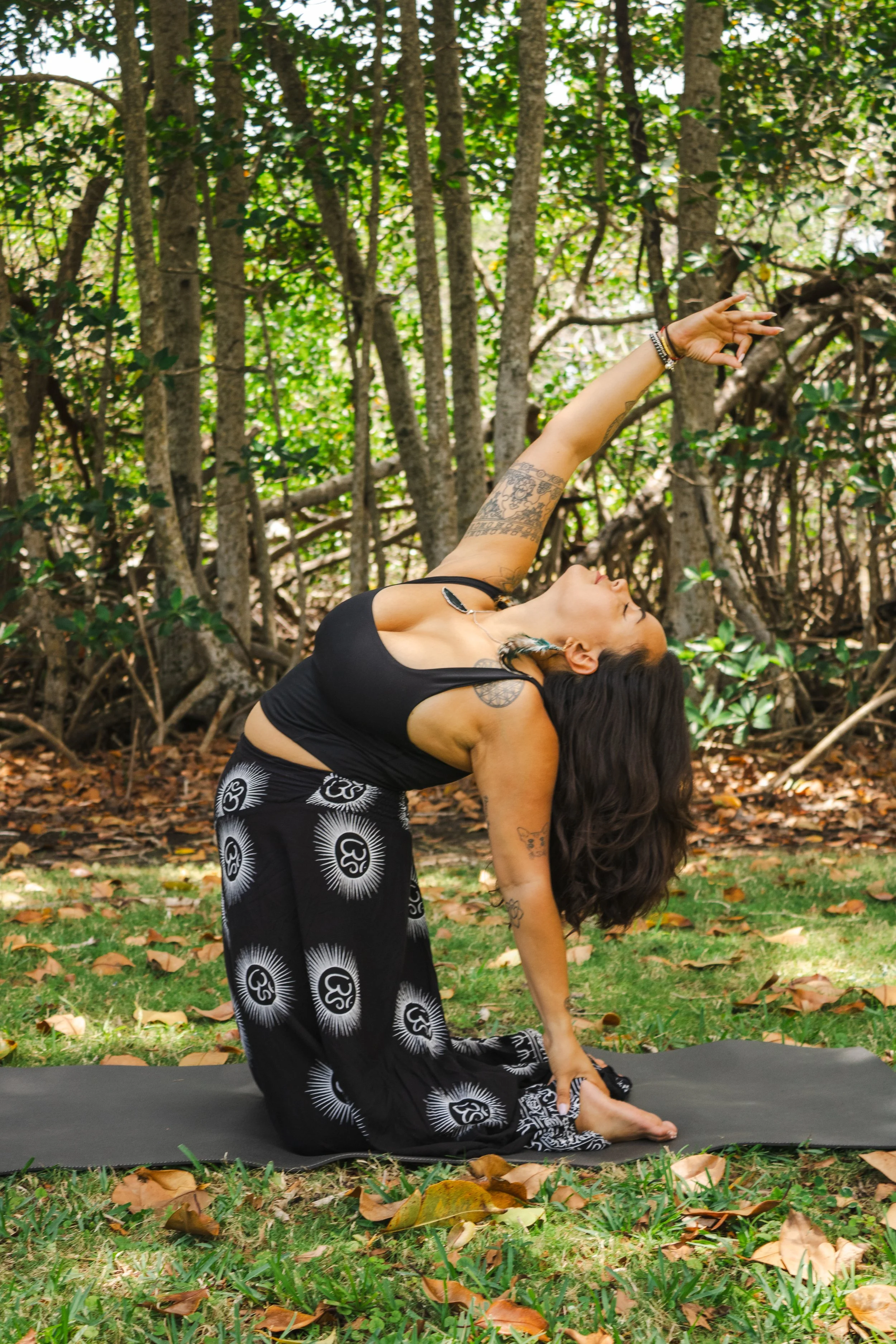A woman practicing yoga outdoors in a wooded area, wearing black patterned pants and a black top, performing a lunge stretch with one arm reaching back and the other reaching forward, surrounded by trees and fallen leaves.