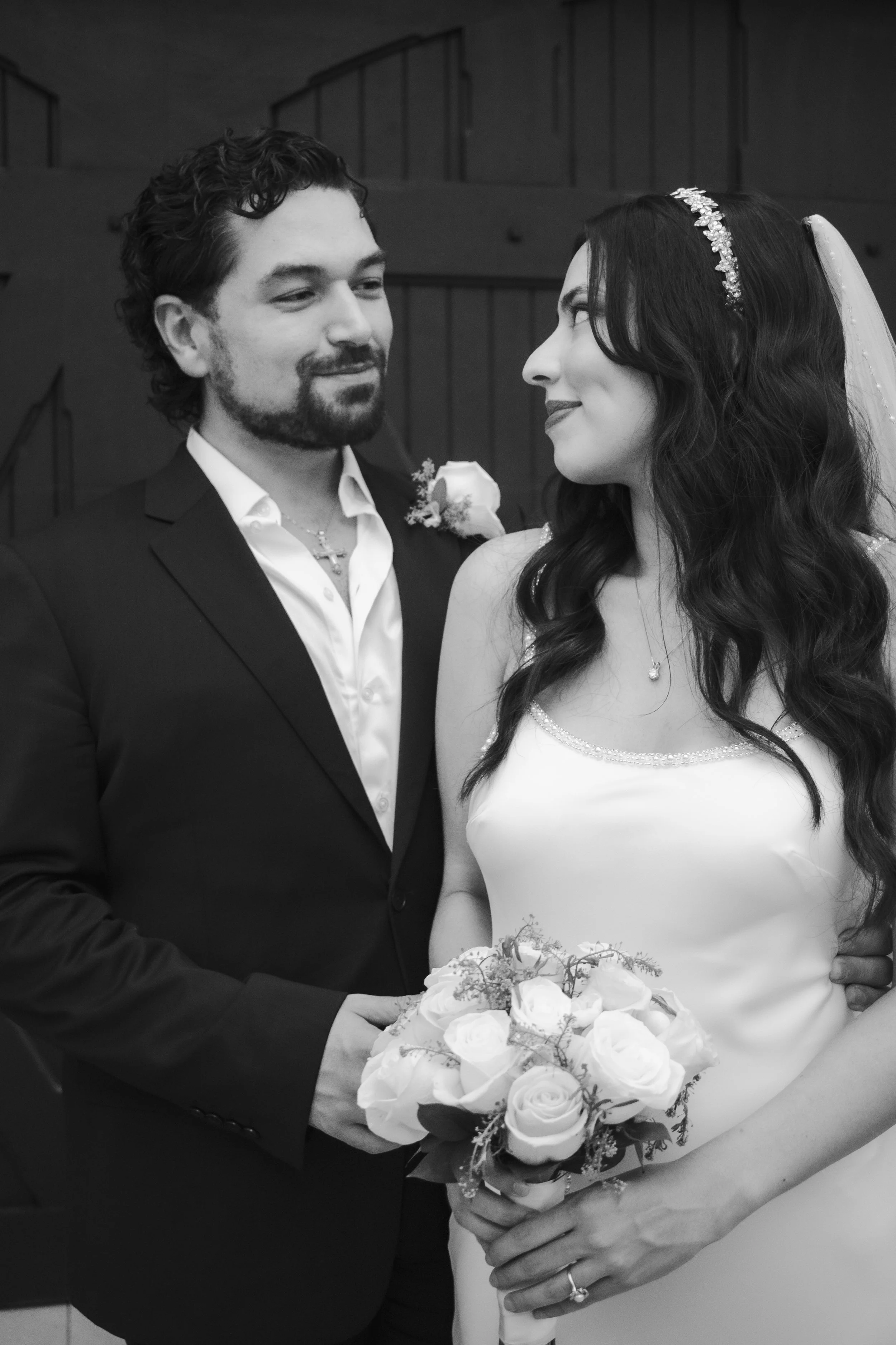 A bride and groom at their wedding, standing close and looking at each other. The groom wears a dark suit and white shirt, the bride wears a white wedding dress with a necklace and headband. They hold a bouquet of roses together.