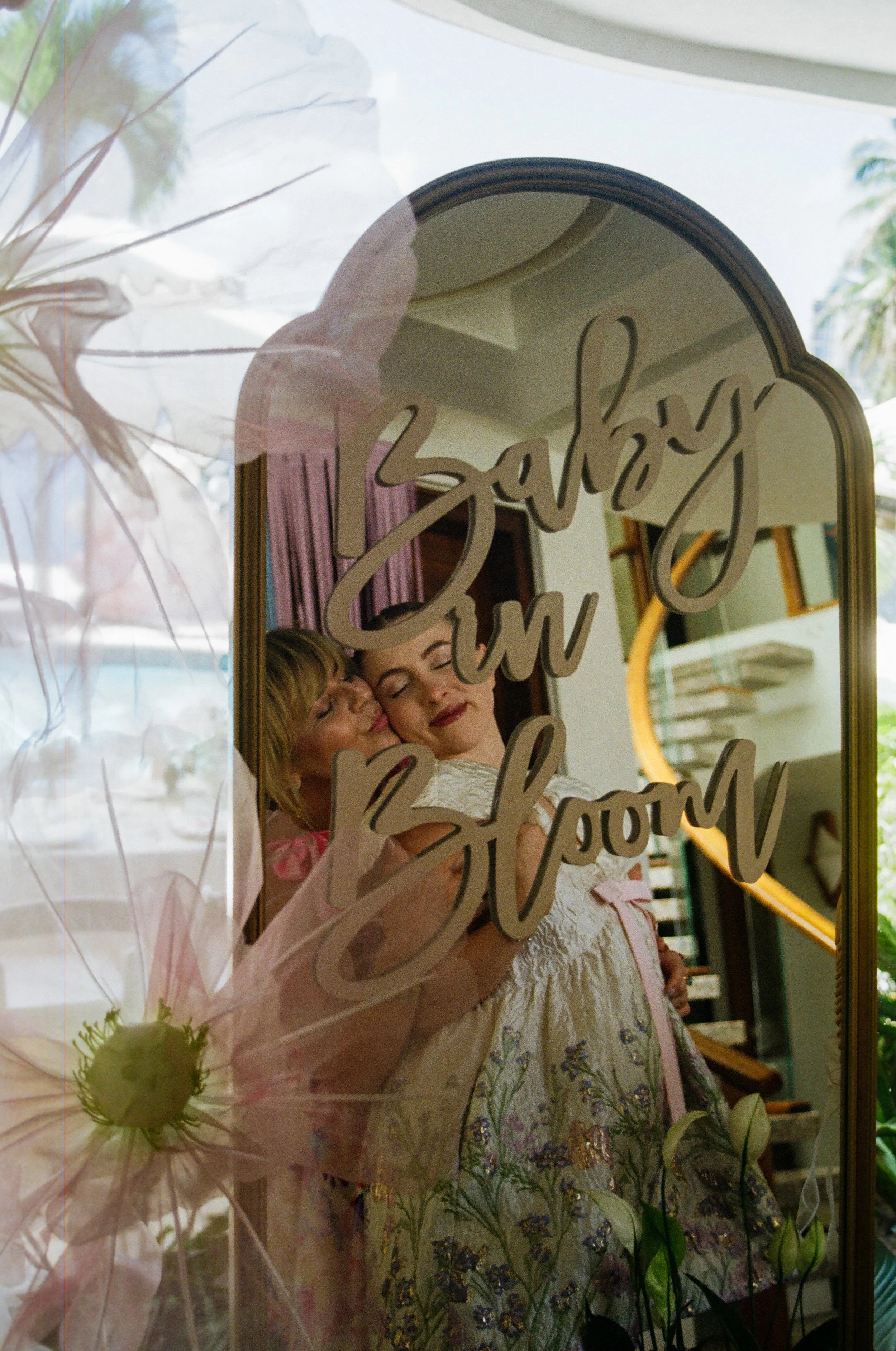 A mirror with a decorative text saying 'you're loved' reflecting two women hugging, surrounded by flowers and greenery, with a room in the background.