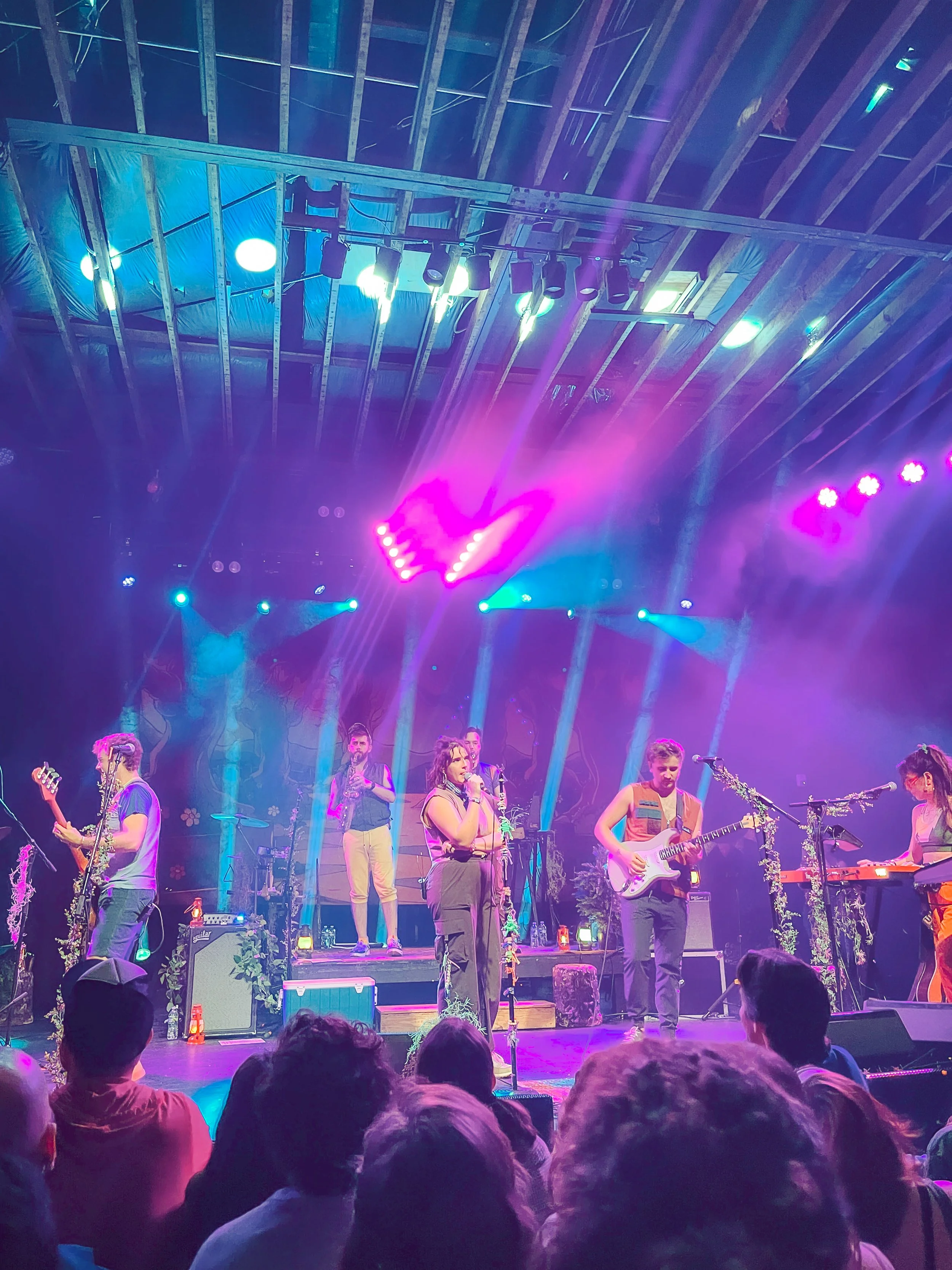 A live music performance on stage with several musicians, colorful lighting including pink and blue lights, and an audience watching.