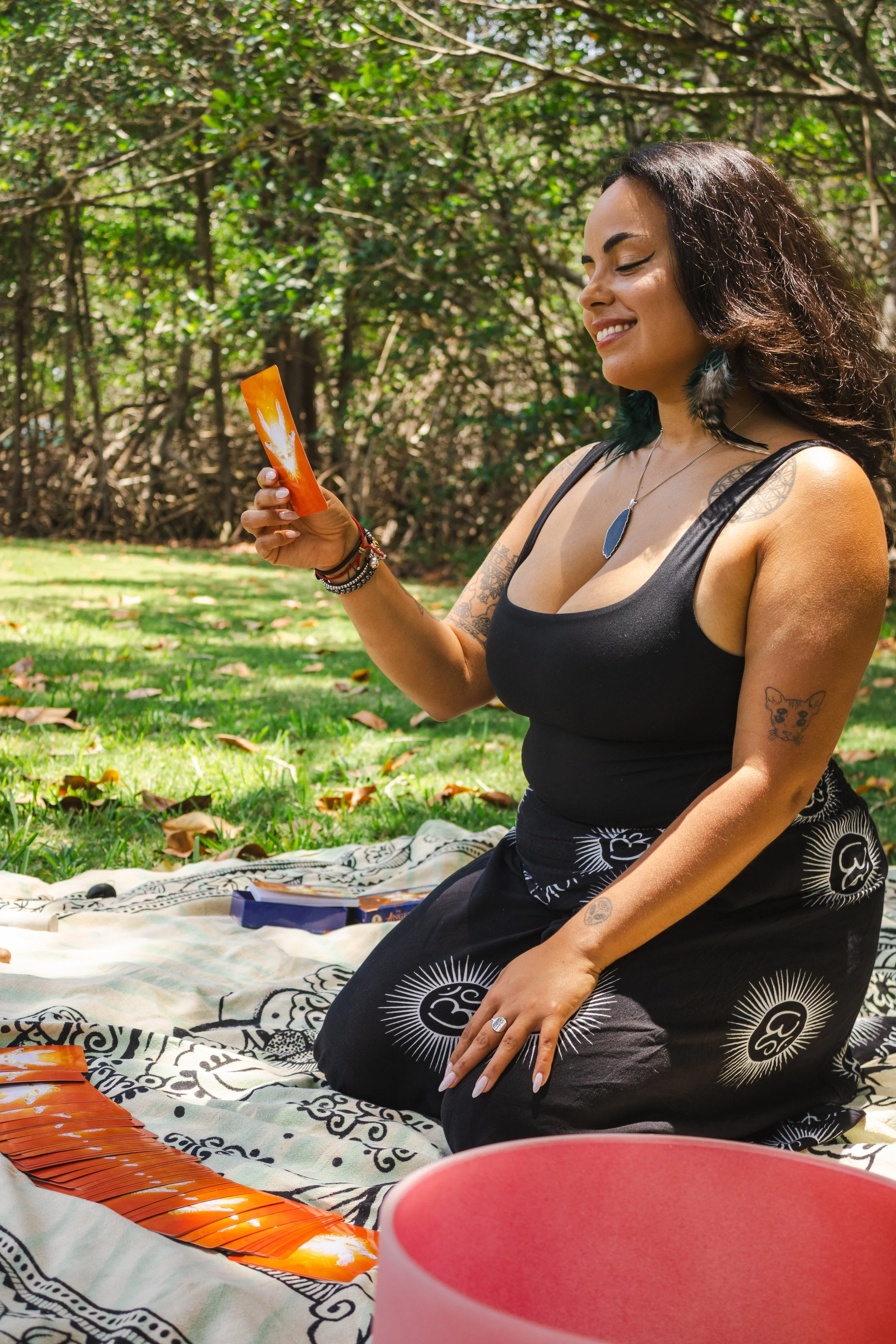 A woman with tattoos on her arm and shoulder, wearing a black tank top, sitting outdoors on a patterned blanket in a wooded area, smiling while holding a bundle of tarot cards.