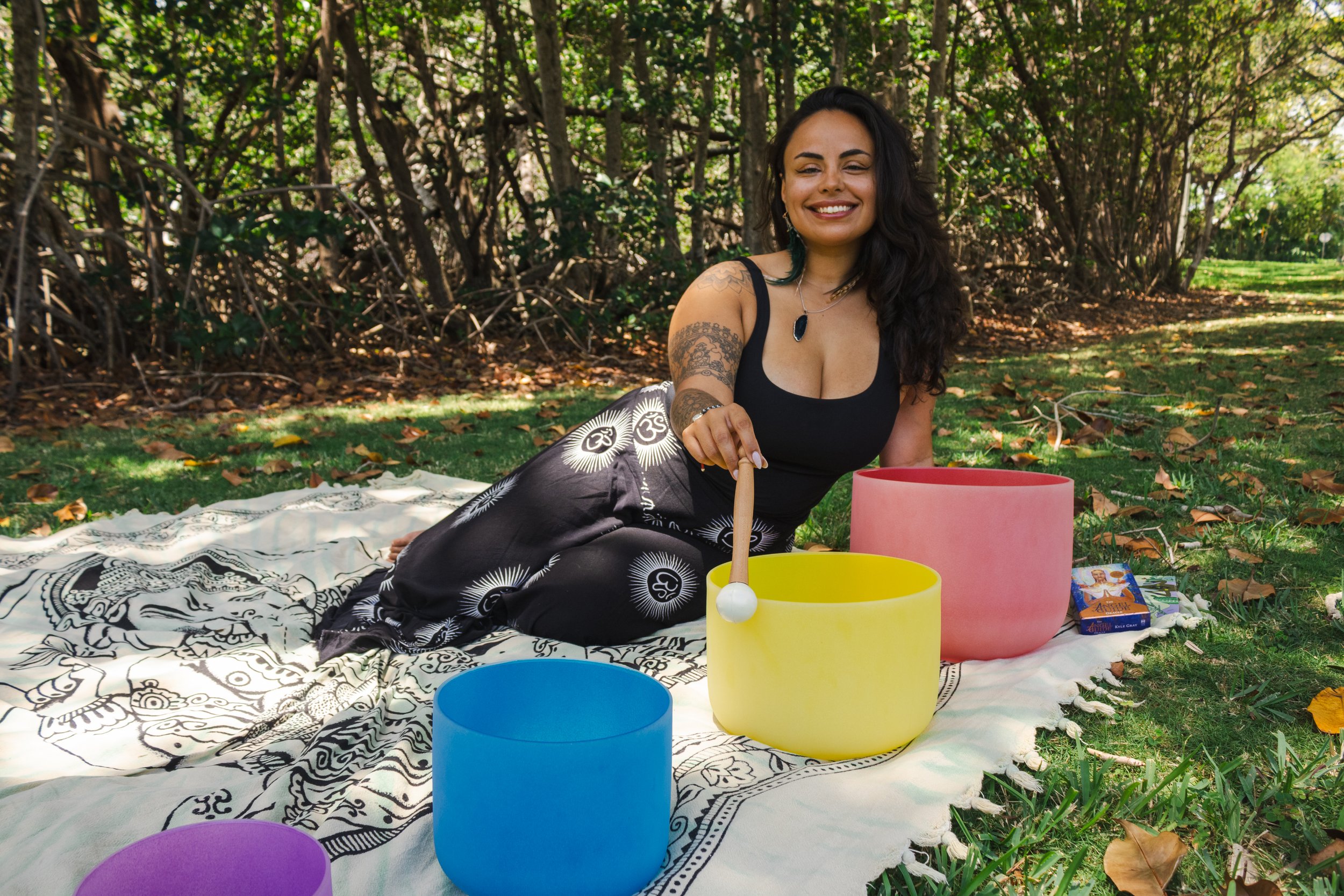 Woman sitting on a blanket in a park, smiling, with colorful singing bowls and a card with a spiritual image nearby.