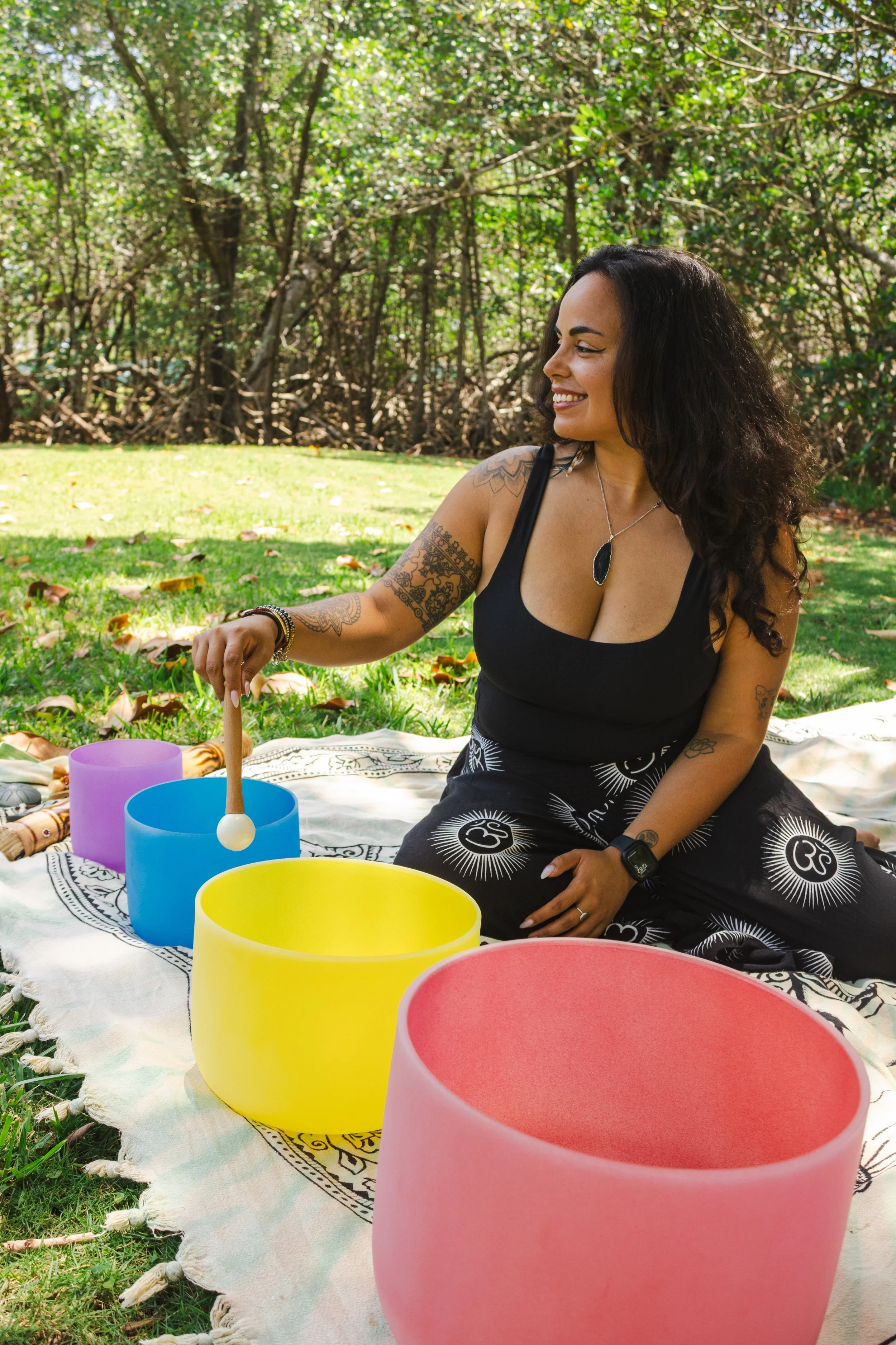A woman with dark curly hair smiling and playing a crystal singing bowl outdoors during daytime. She is wearing a black tank top and patterned pants, seated on a blanket with other colorful singing bowls in front of her, surrounded by green grass and