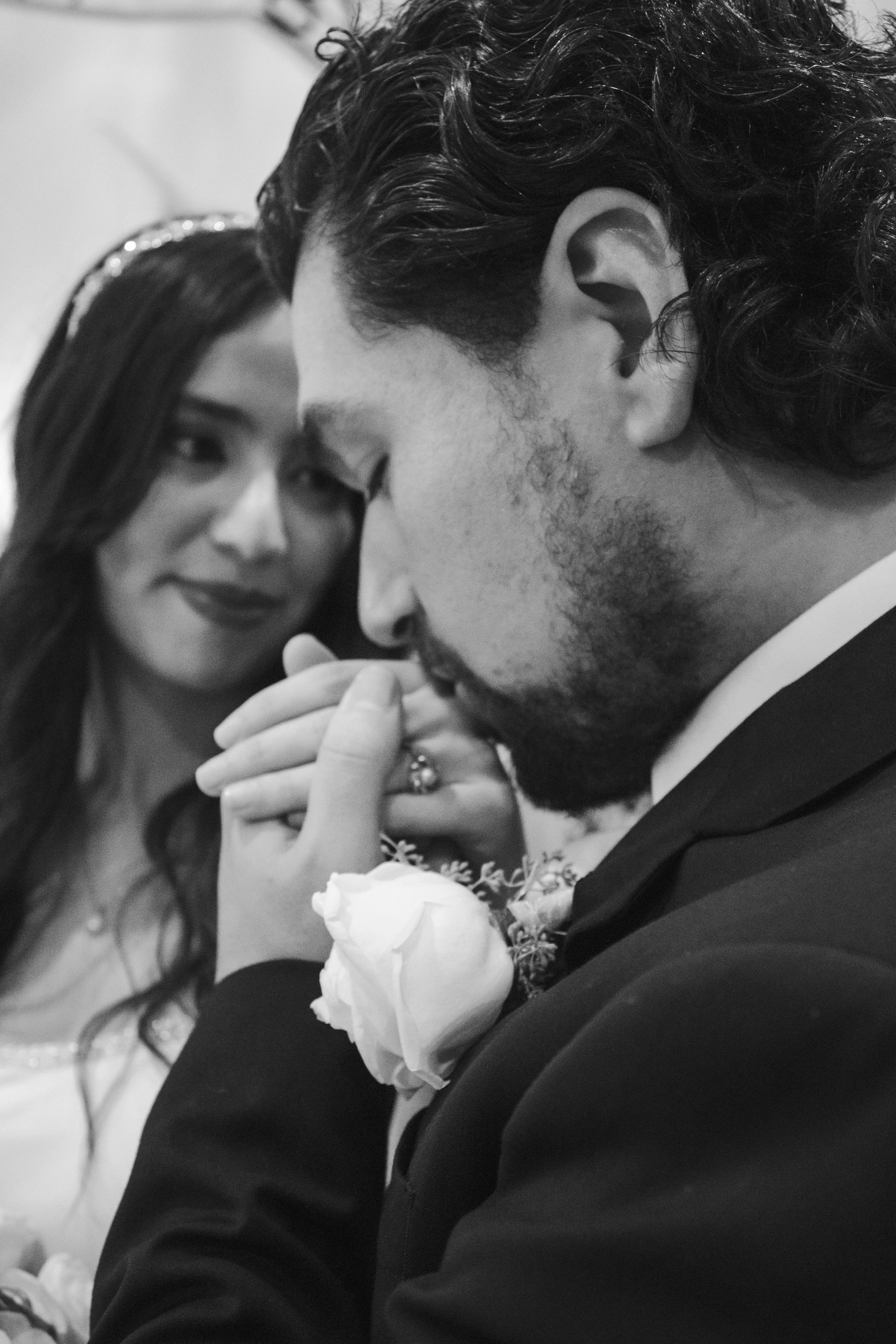 Black and white photo of a woman and man in a close moment. The woman is smiling softly, while the man is in the foreground, wearing a suit and boutonniere, holding a white rose. The woman appears to be touching his face or neck.