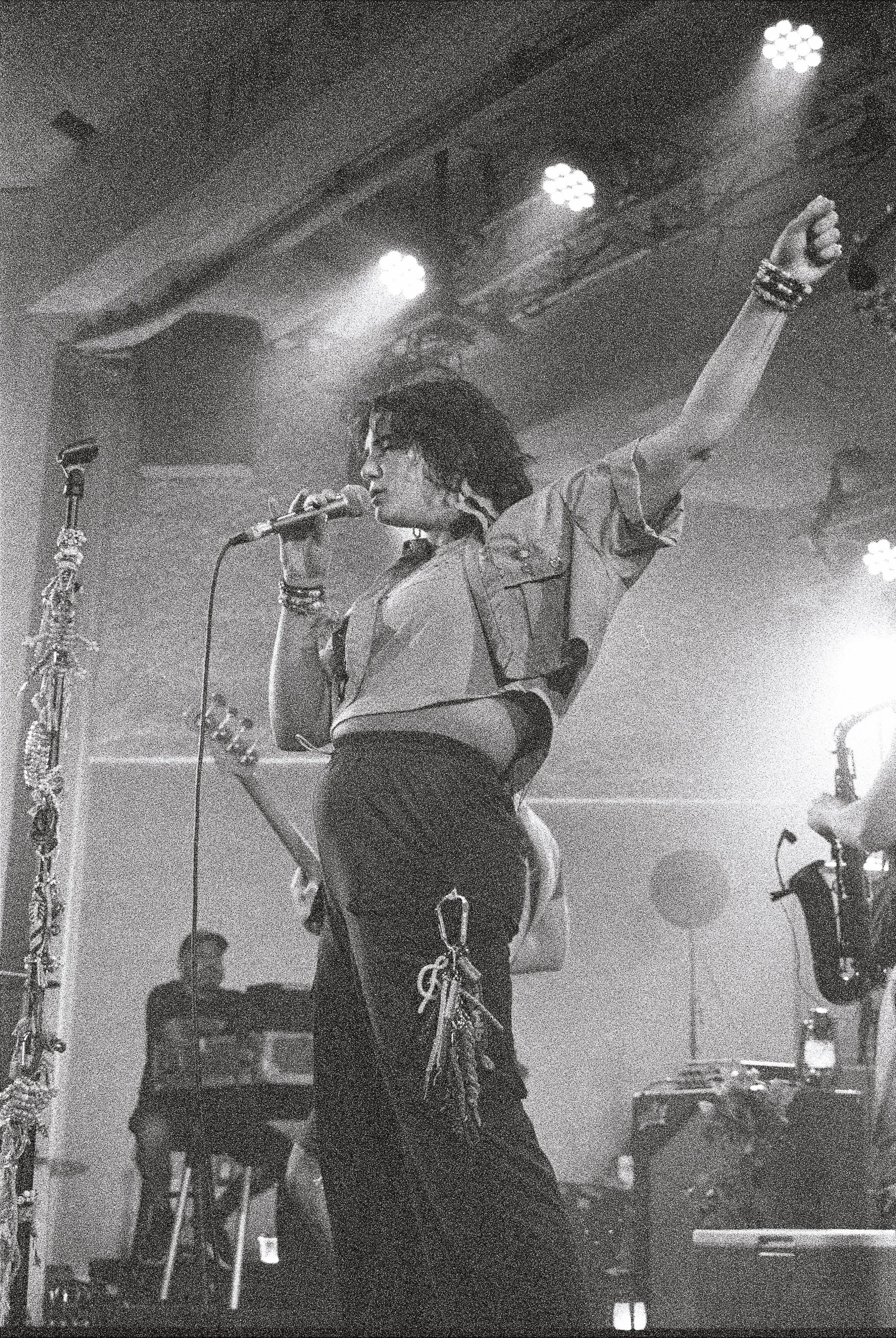 A woman singing into a microphone on stage with her right arm raised, wearing sunglasses, a short-sleeve shirt, and jewelry. There are stage lights overhead.