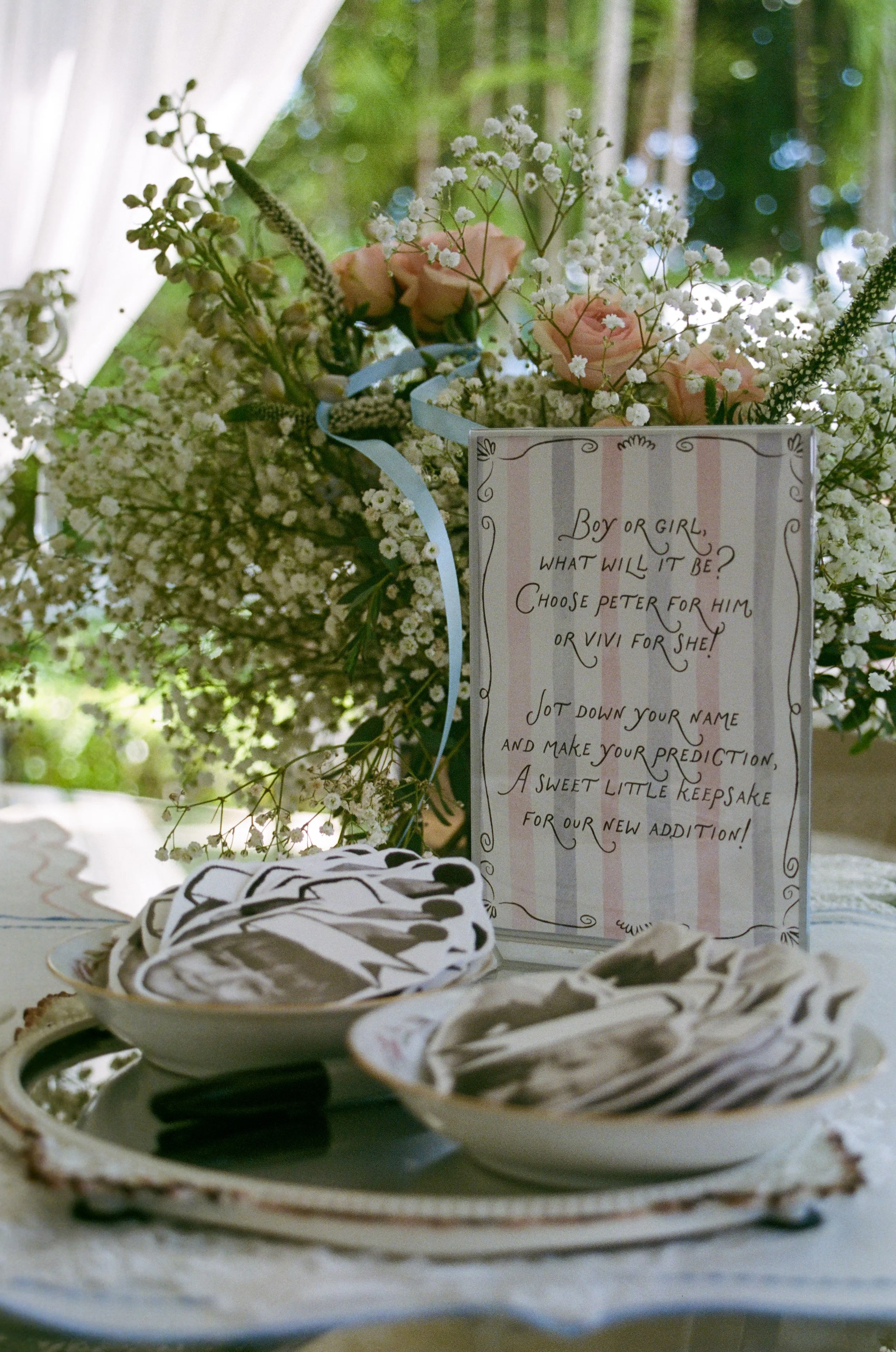 A table with a floral arrangement of pink roses, white baby's breath, and greenery, and a handwritten sign asking guests to jot down their names and predictions for a couple's new addition. There are plates of cookies in the foreground.