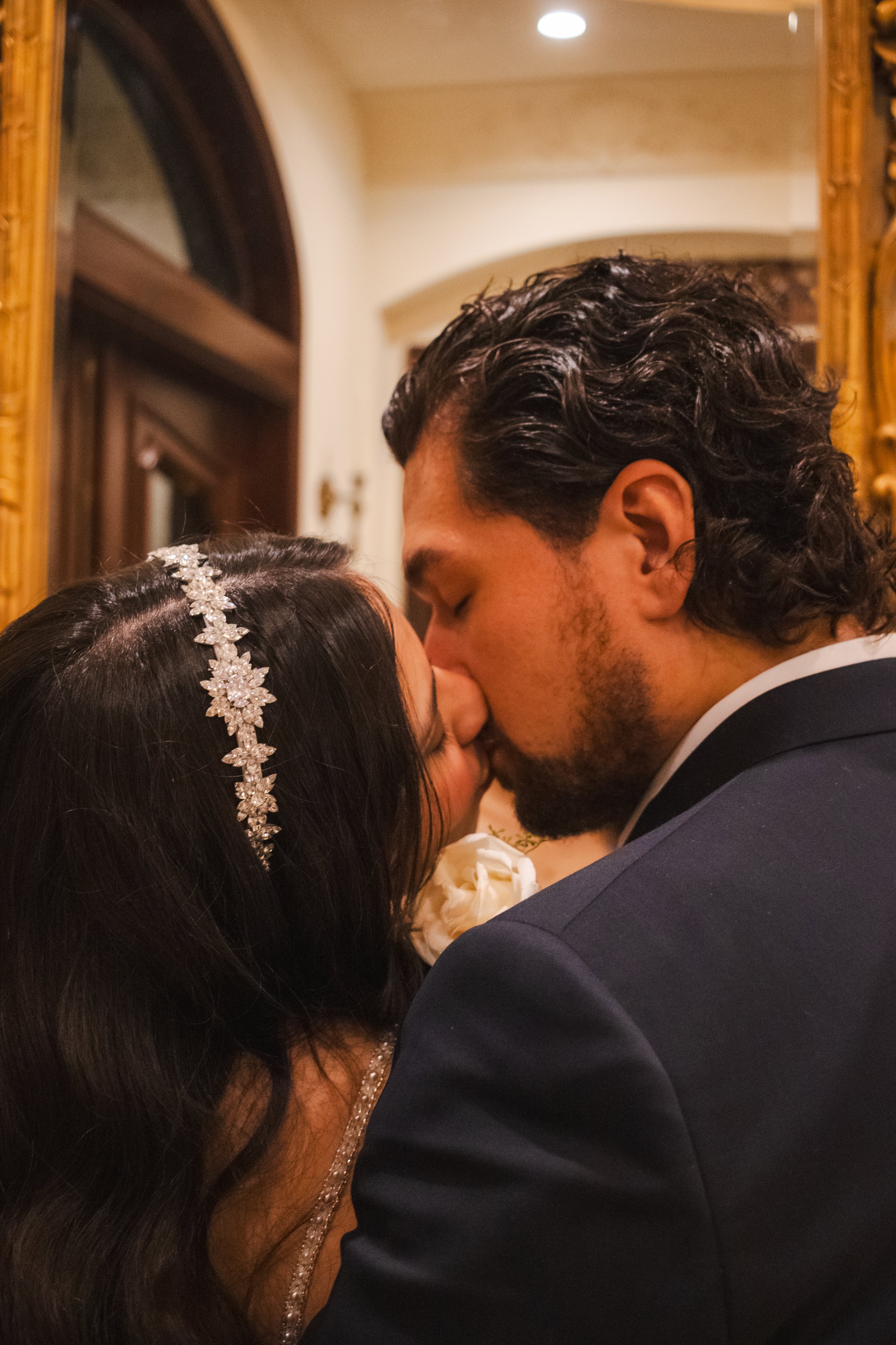 A bride and groom are sharing a kiss at their wedding, with the bride wearing a decorative headband and the groom in a dark tuxedo, inside a warmly lit room with reflection in a mirror.