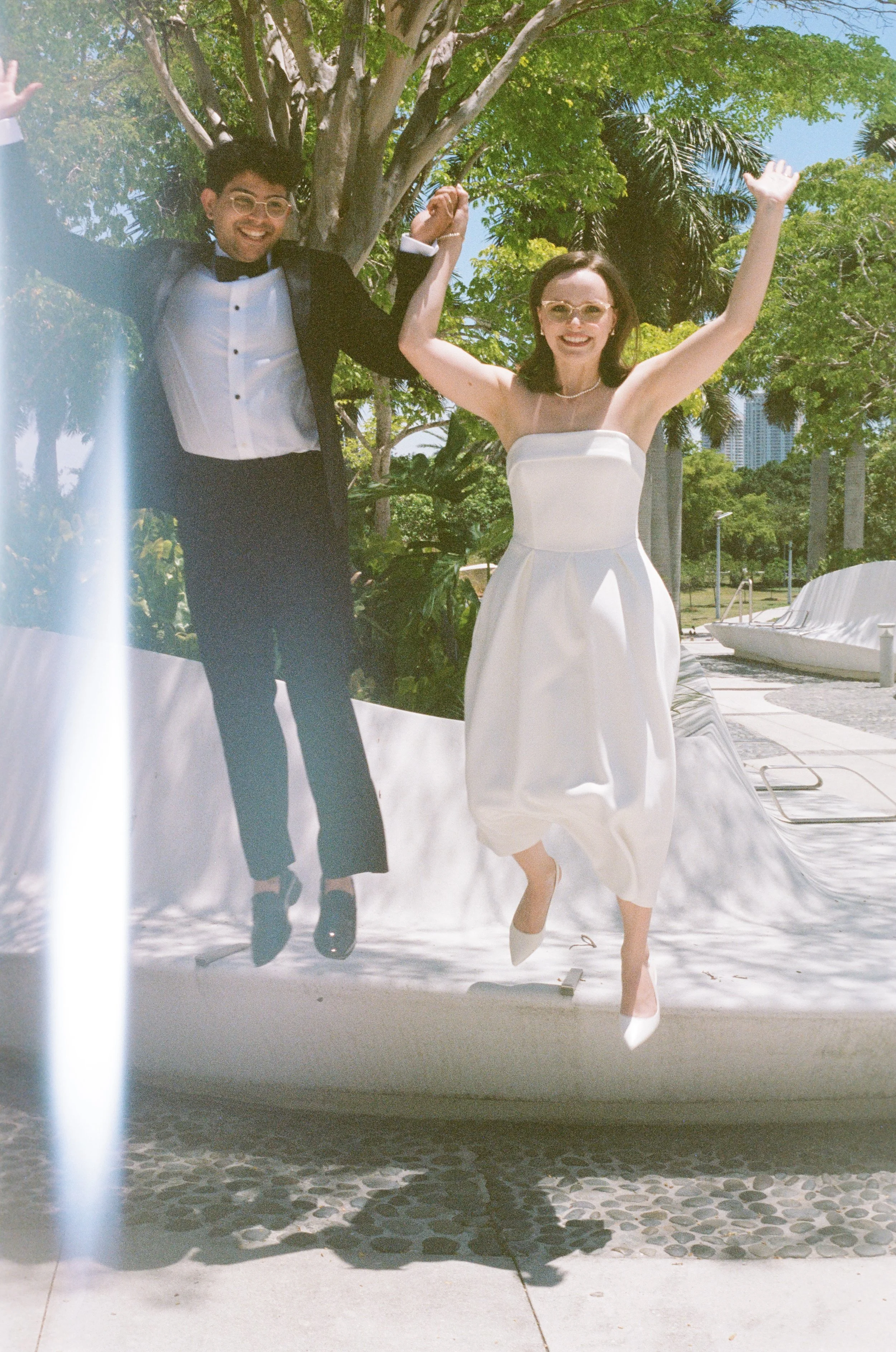 A man and woman at a wedding, mid-air, with trees and a cityscape in the background. The man is in a tuxedo and the woman in a white wedding dress, both smiling.