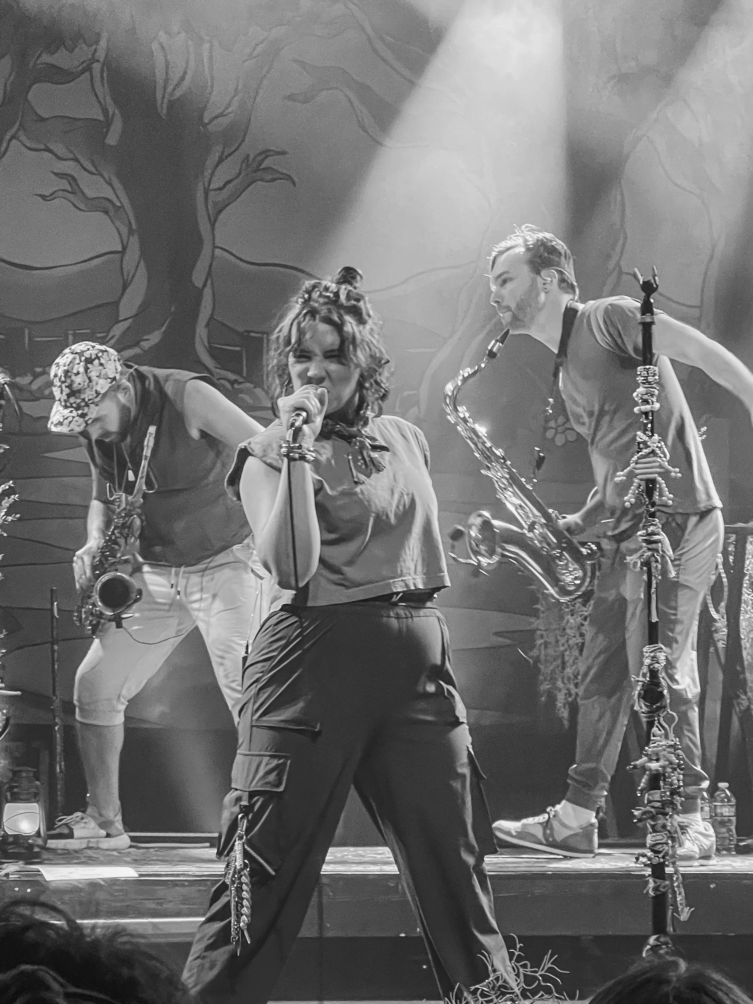 Black and white photo of a musical performance with a woman singing into a microphone and a man playing a saxophone, in front of a backdrop with a large tree design.