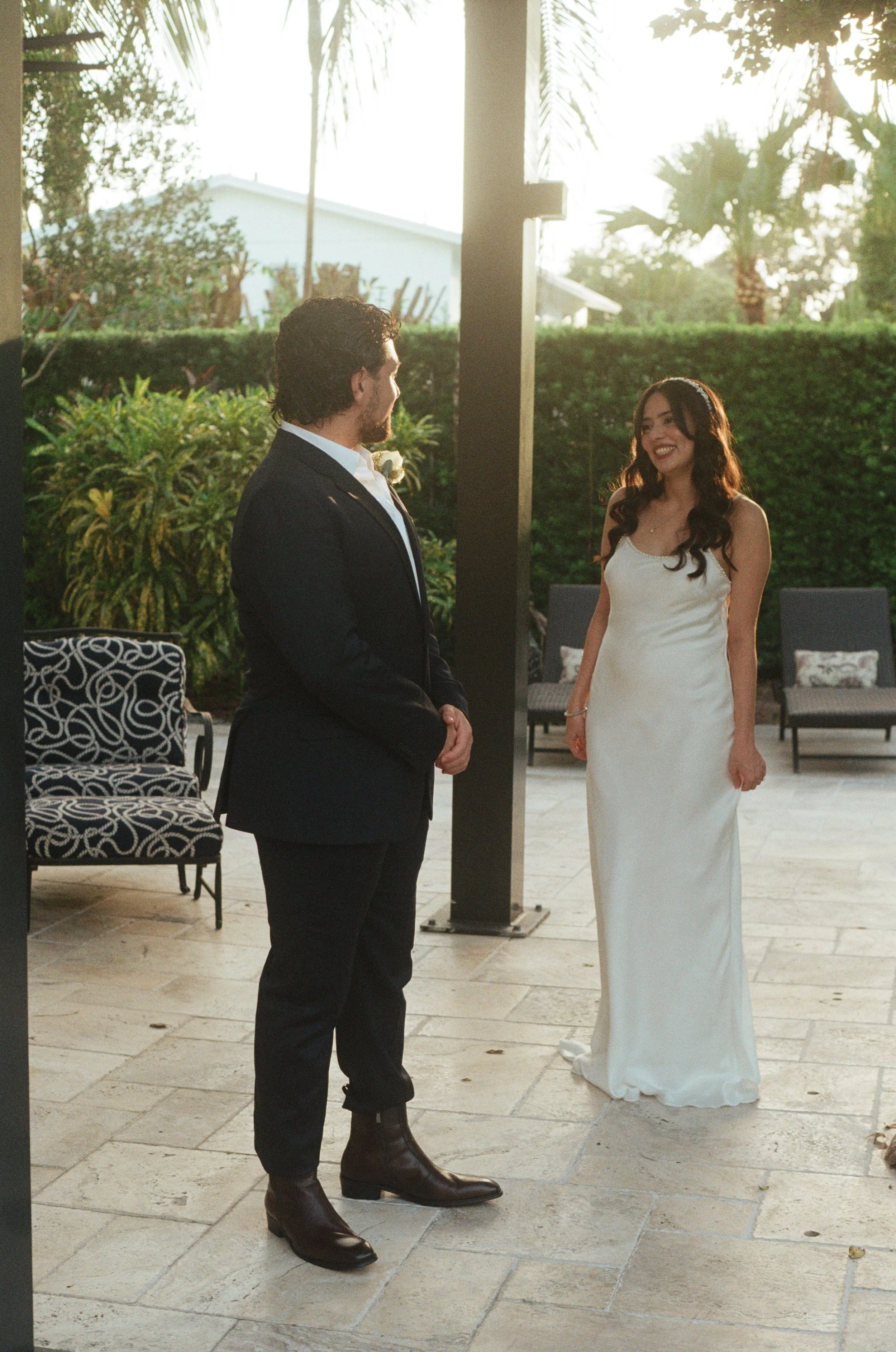 A man and woman are standing outdoors, facing each other. The man is dressed in a black suit, white shirt, and black boots, and the woman is wearing a white sleeveless dress. They appear to be smiling and engaging in conversation.