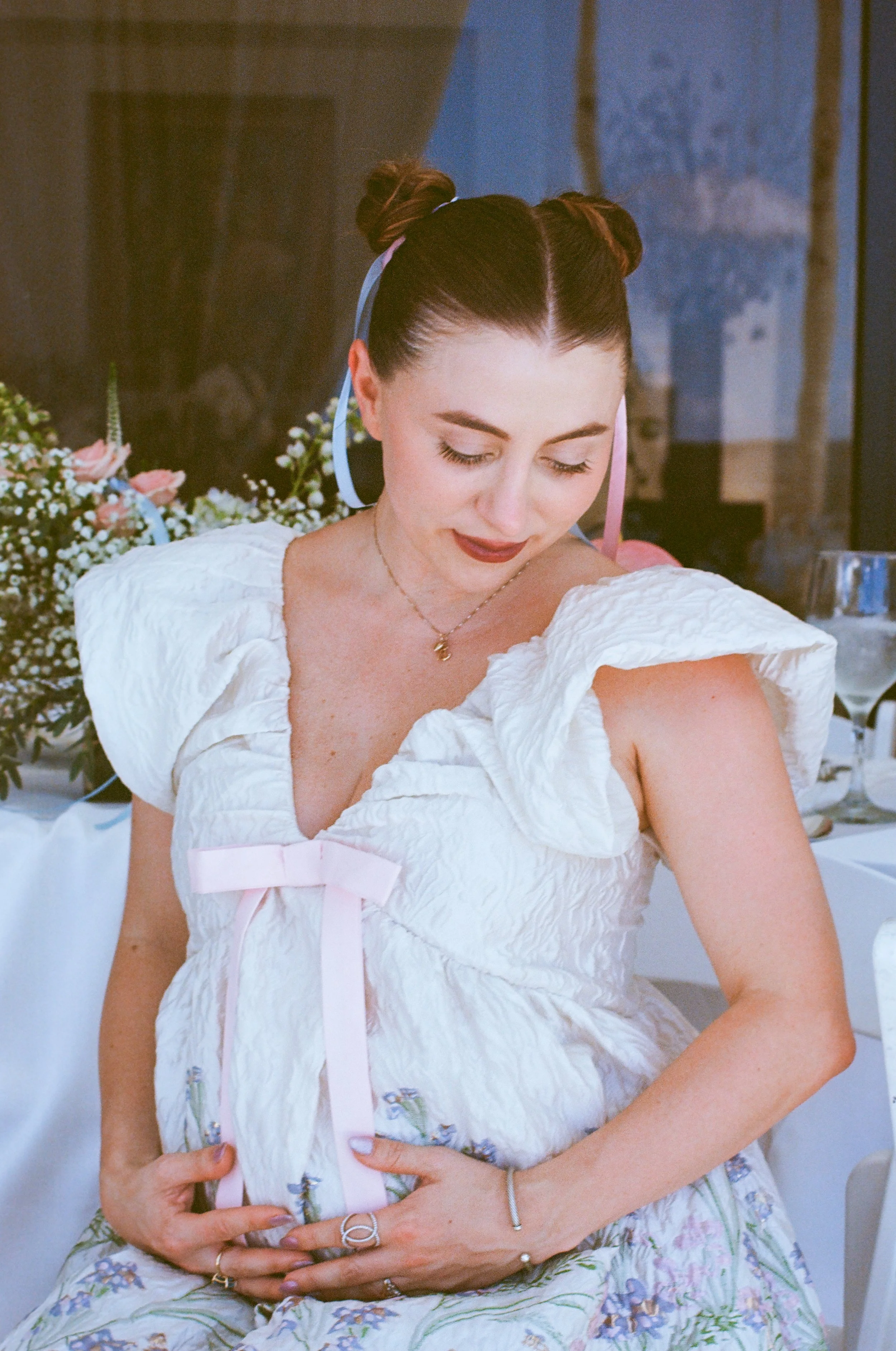 A woman with dark hair styled in two buns, wearing a white dress with puffy sleeves and a pink ribbon, is sitting indoors with a bouquet of flowers beside her. She is looking down and smiling softly, with jewelry including rings, a bracelet, and a ne
