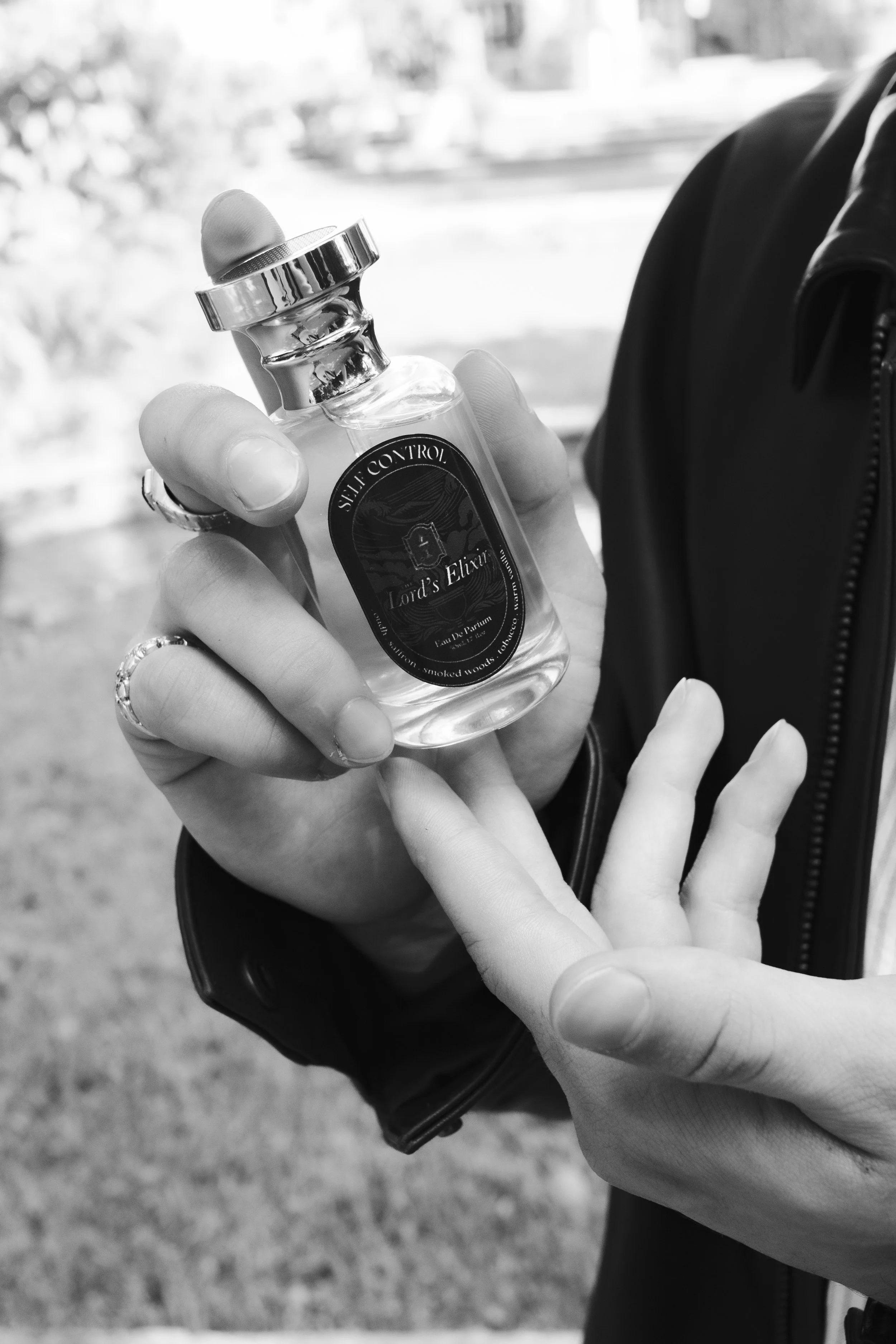 Close-up of a hand holding a small perfume bottle labeled 'Lord's Elixir' with a metallic cap and a ring on the finger. The background is blurred outside view.