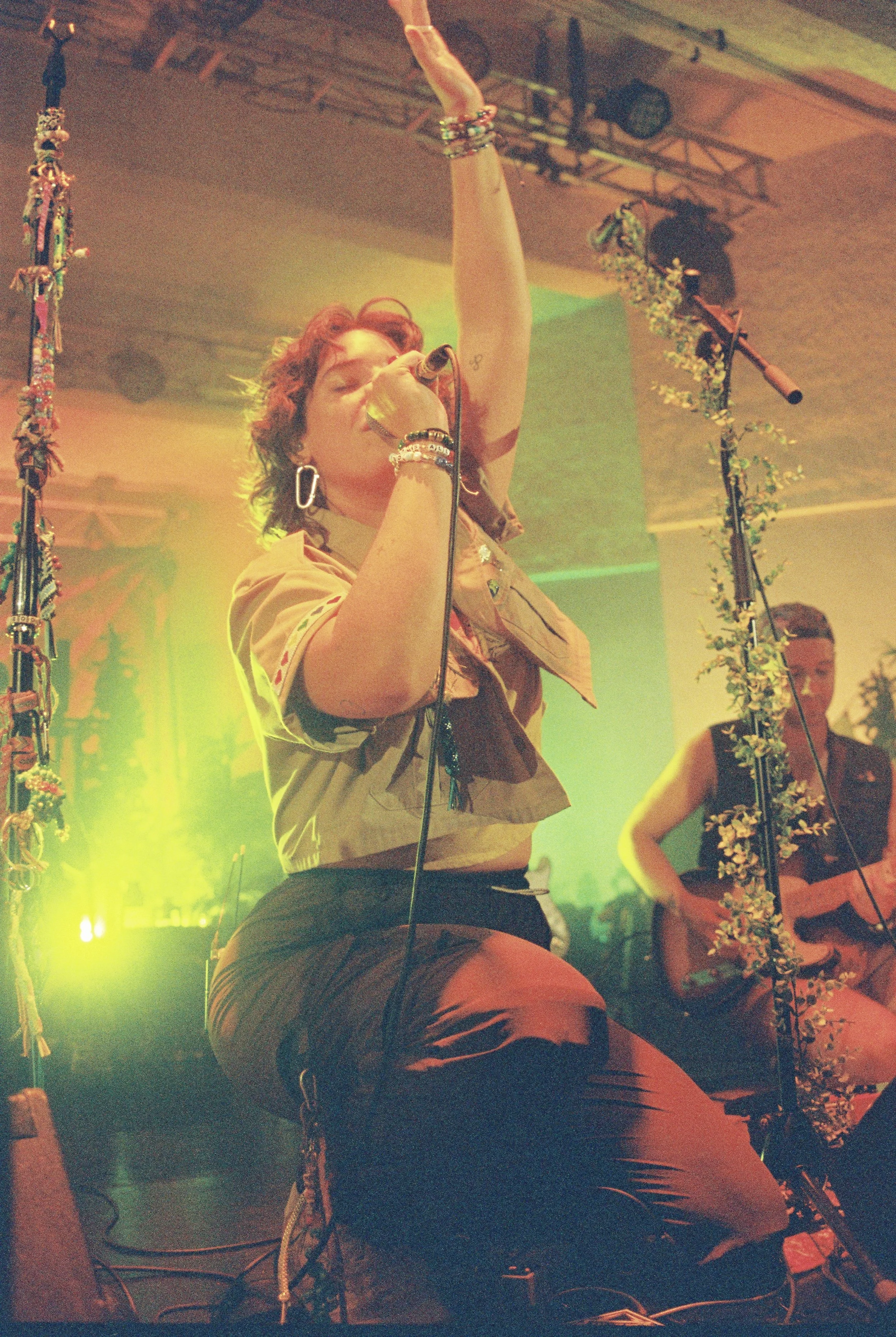 A woman singing into a microphone on stage, wearing a tan vest, black shorts, and bracelets, with colorful stage lighting and decorative props, including a micstand decorated with flowers to her right.