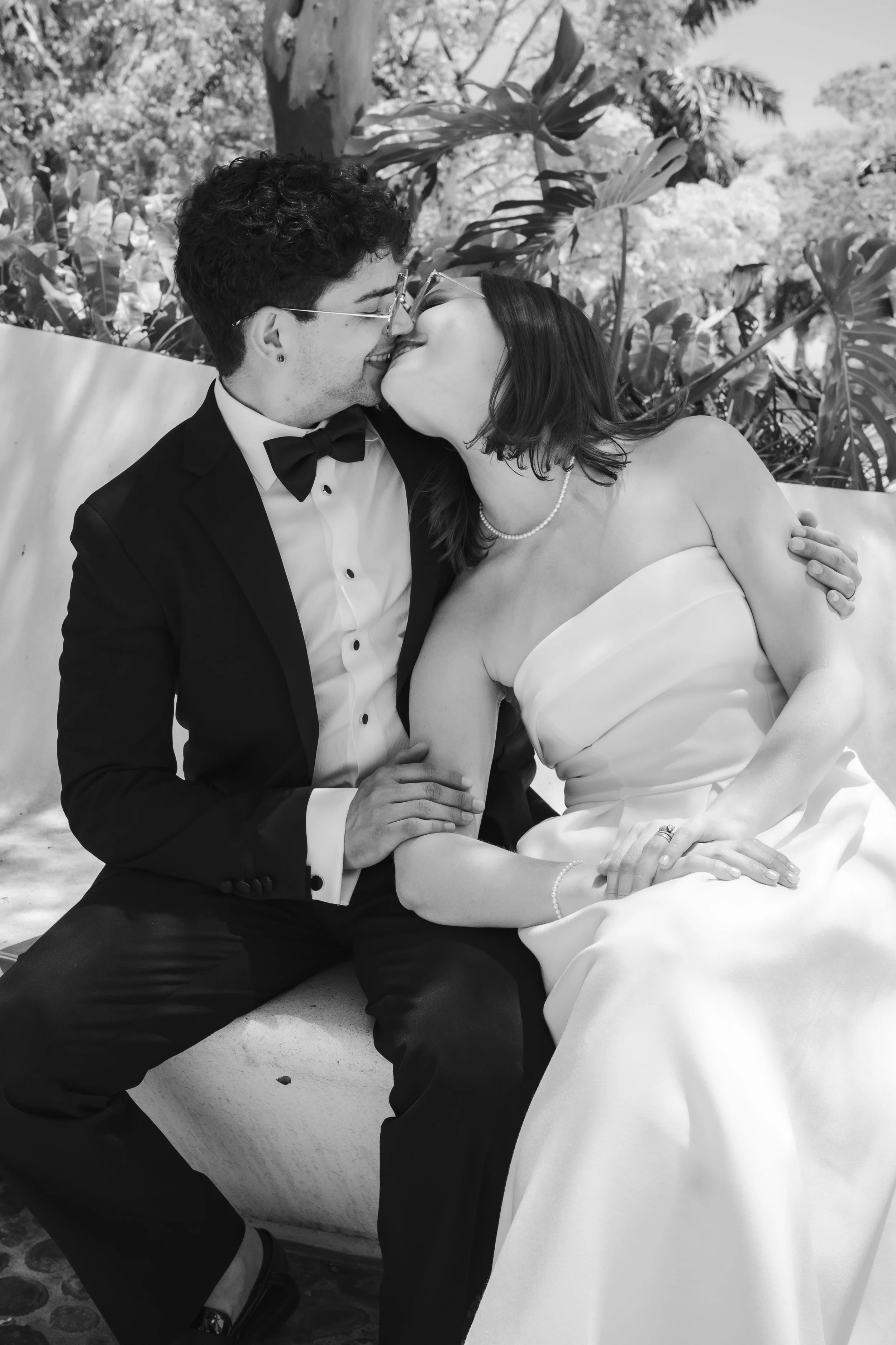 A couple dressed in formal wedding attire sharing a kiss while sitting outdoors in front of lush tropical plants.