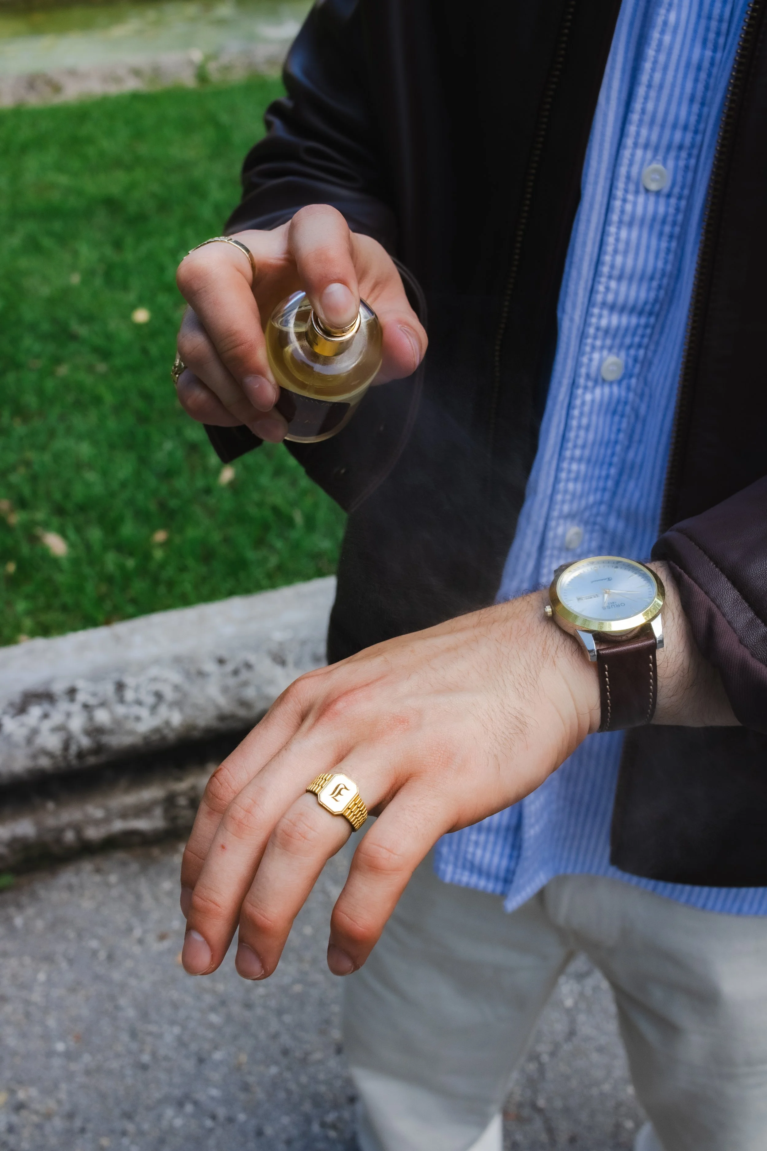 Close-up of two hands with multiple rings and watches, showing off jewelry, outdoors near a grassy area and a stone ledge.