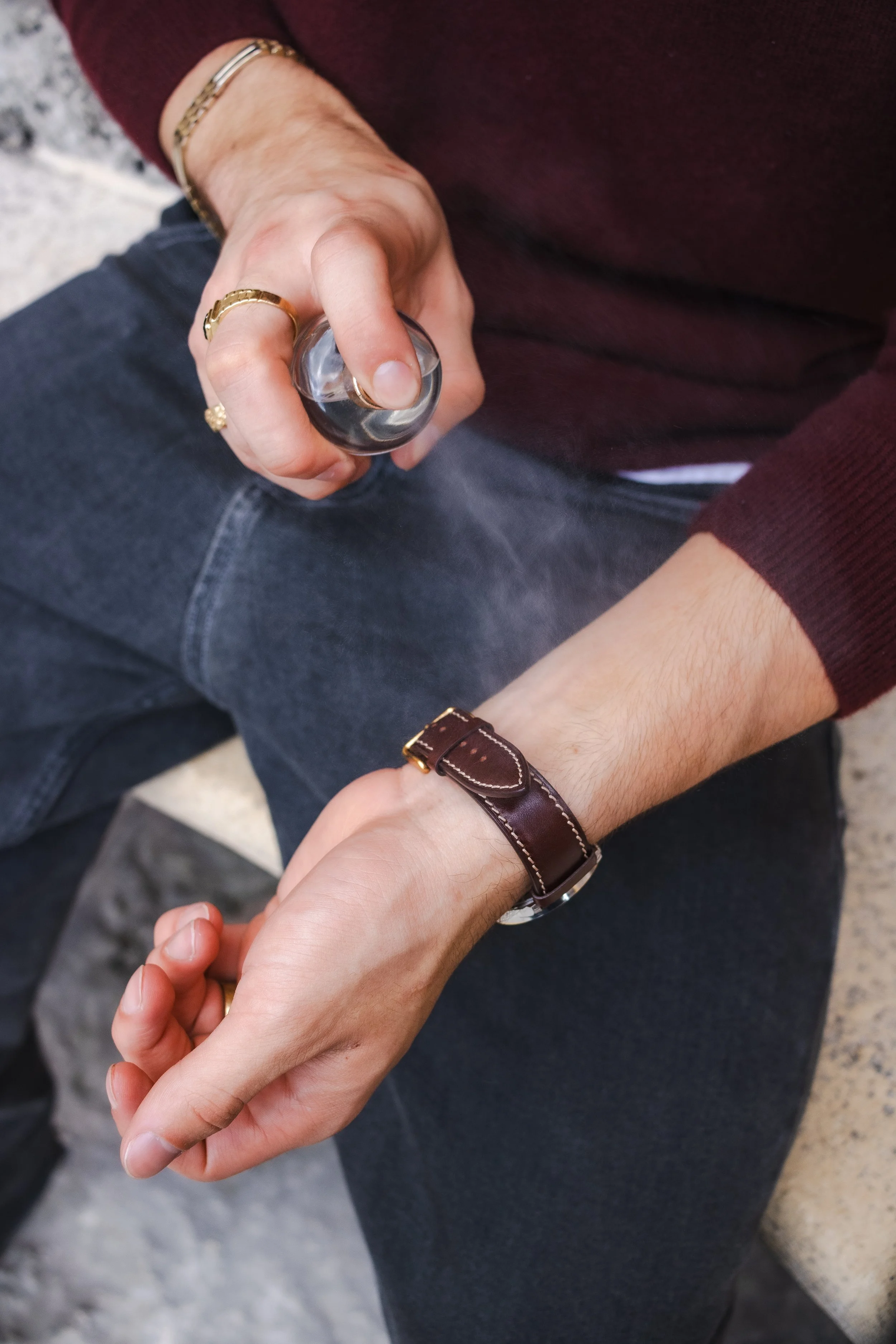 A person is holding a perfume bottle with their right hand, while the left hand is out being sprayed by the perfume. The person is wearing a maroon sweater, dark jeans, and accessories including a gold bracelet, rings, and a leather strap watch.